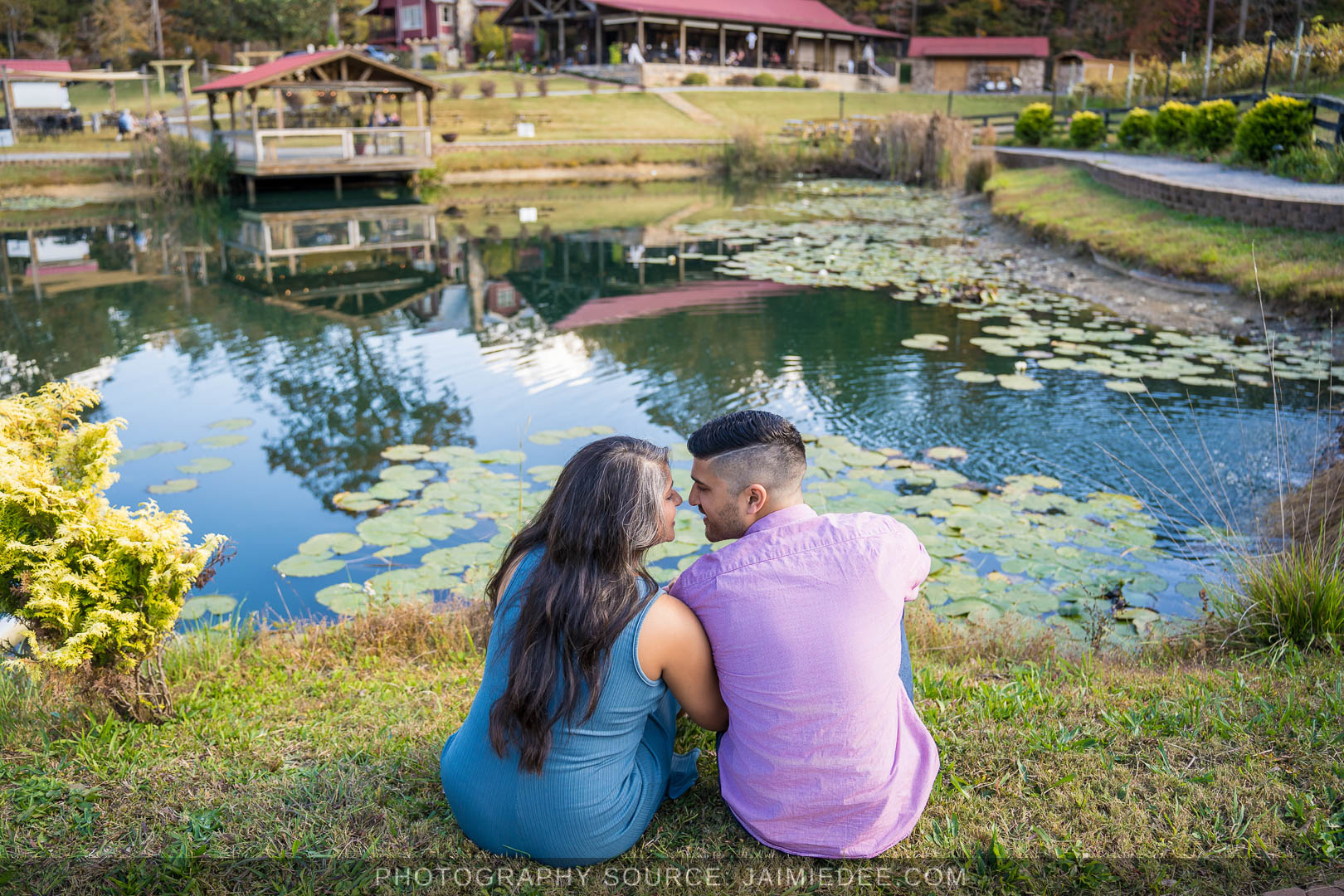Couple sitting on the grassy bank by the lily pads, with the covered pavilion across the lake at Big Door Vineyards