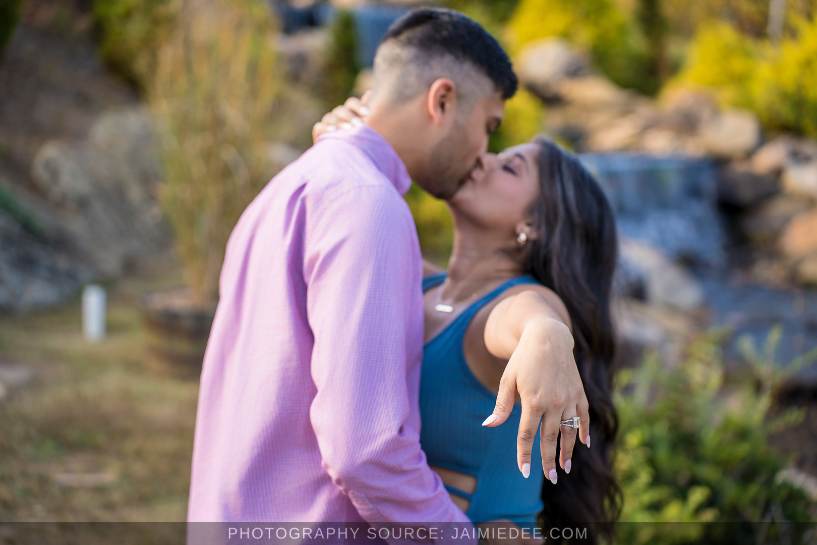 Couple kissing as she shows her engagement ring beside the waterfall at Big Door Vineyards