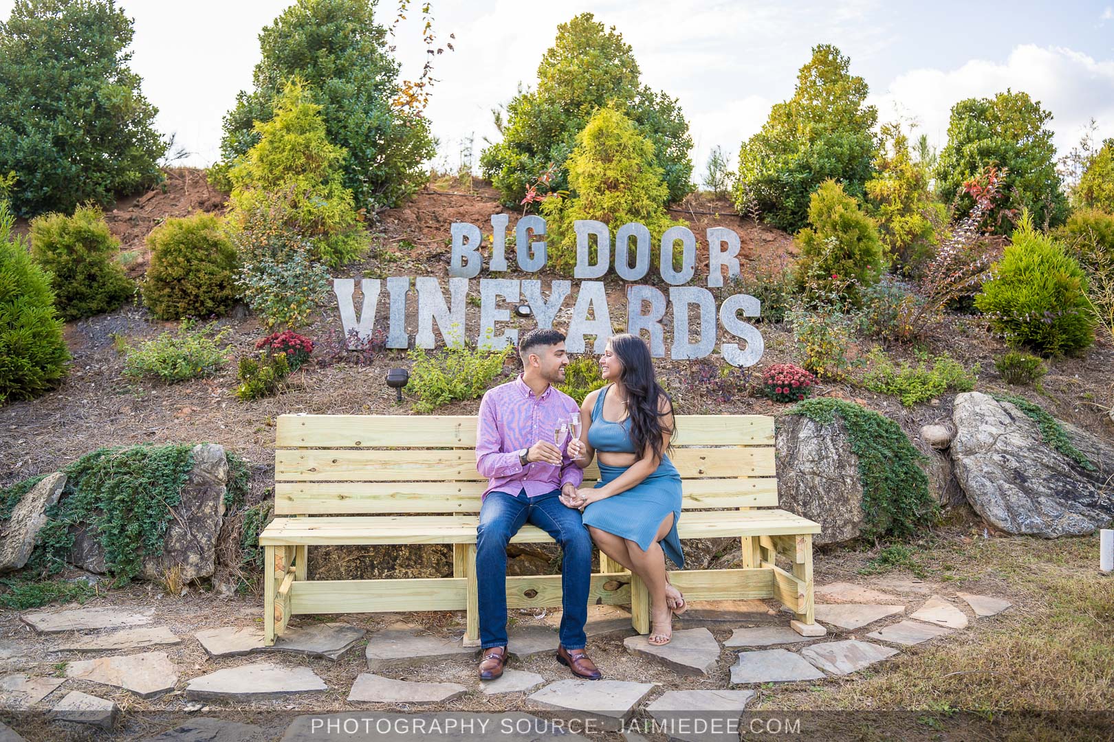 Engaged couple sitting on a bench in front of the Big Door Vineyards sign in White, GA