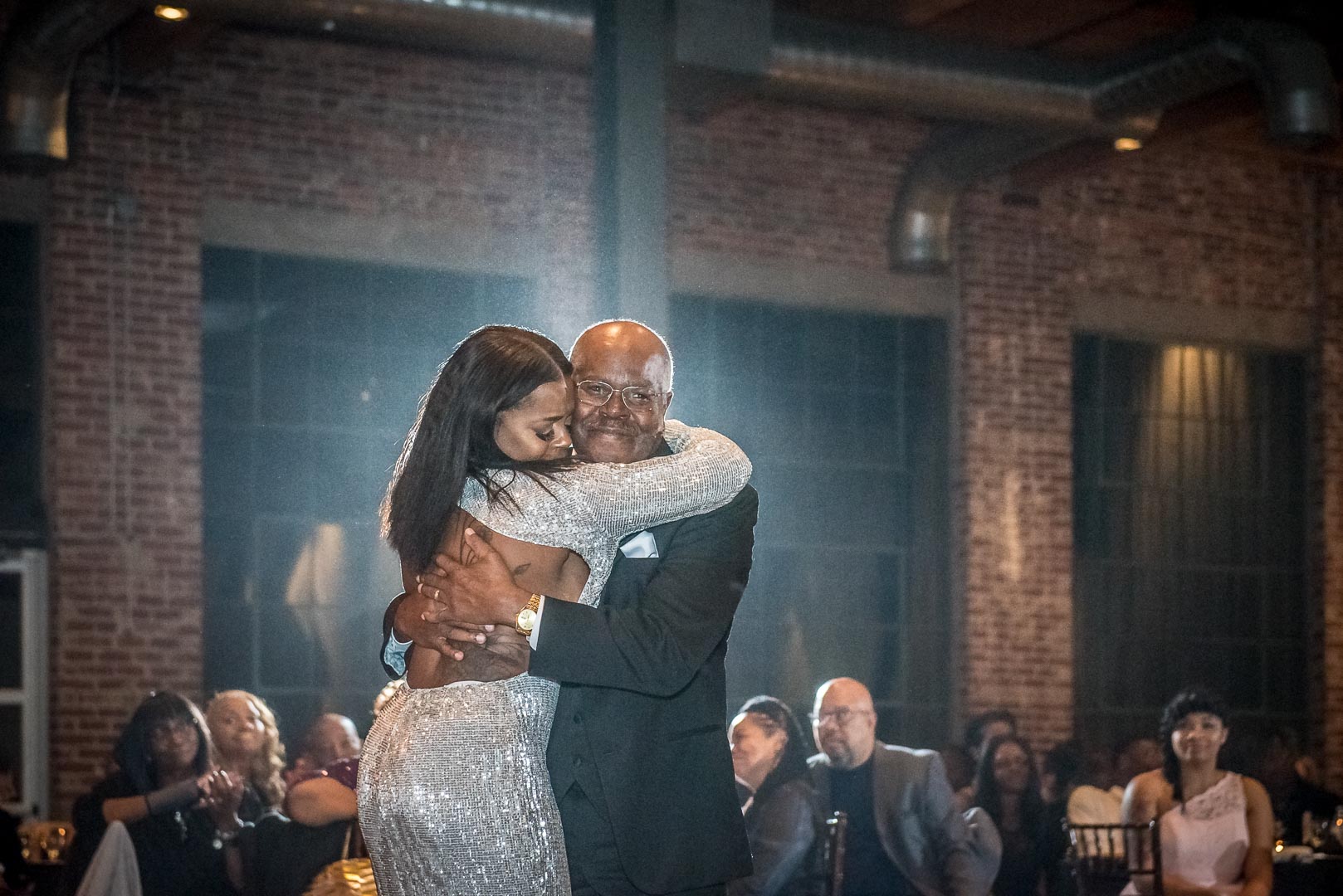 Bride hugging her father during their parent dance in a brick-walled reception hall