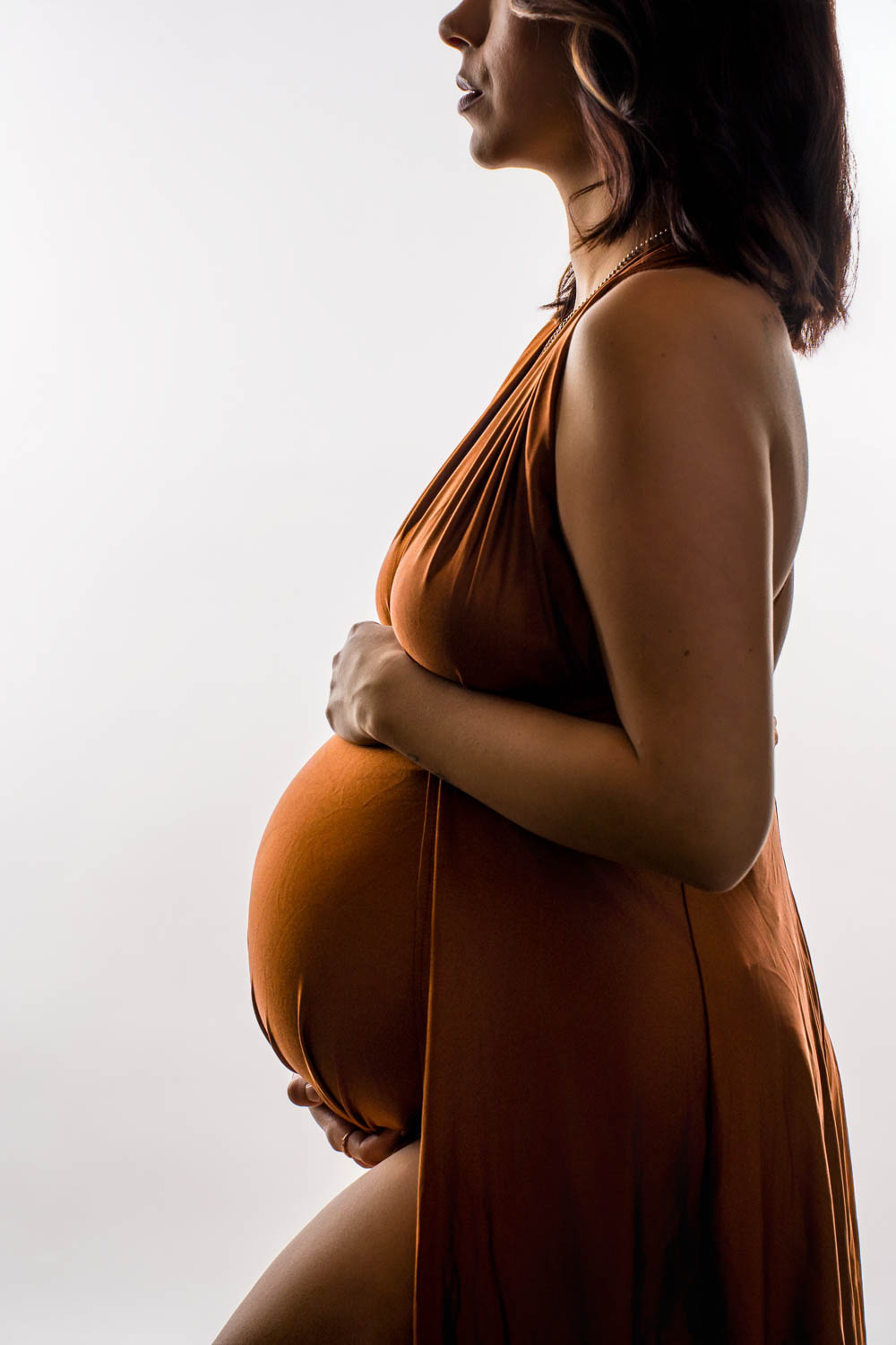 Side profile maternity portrait of an expecting mother in a rust-colored dress against a white backdrop