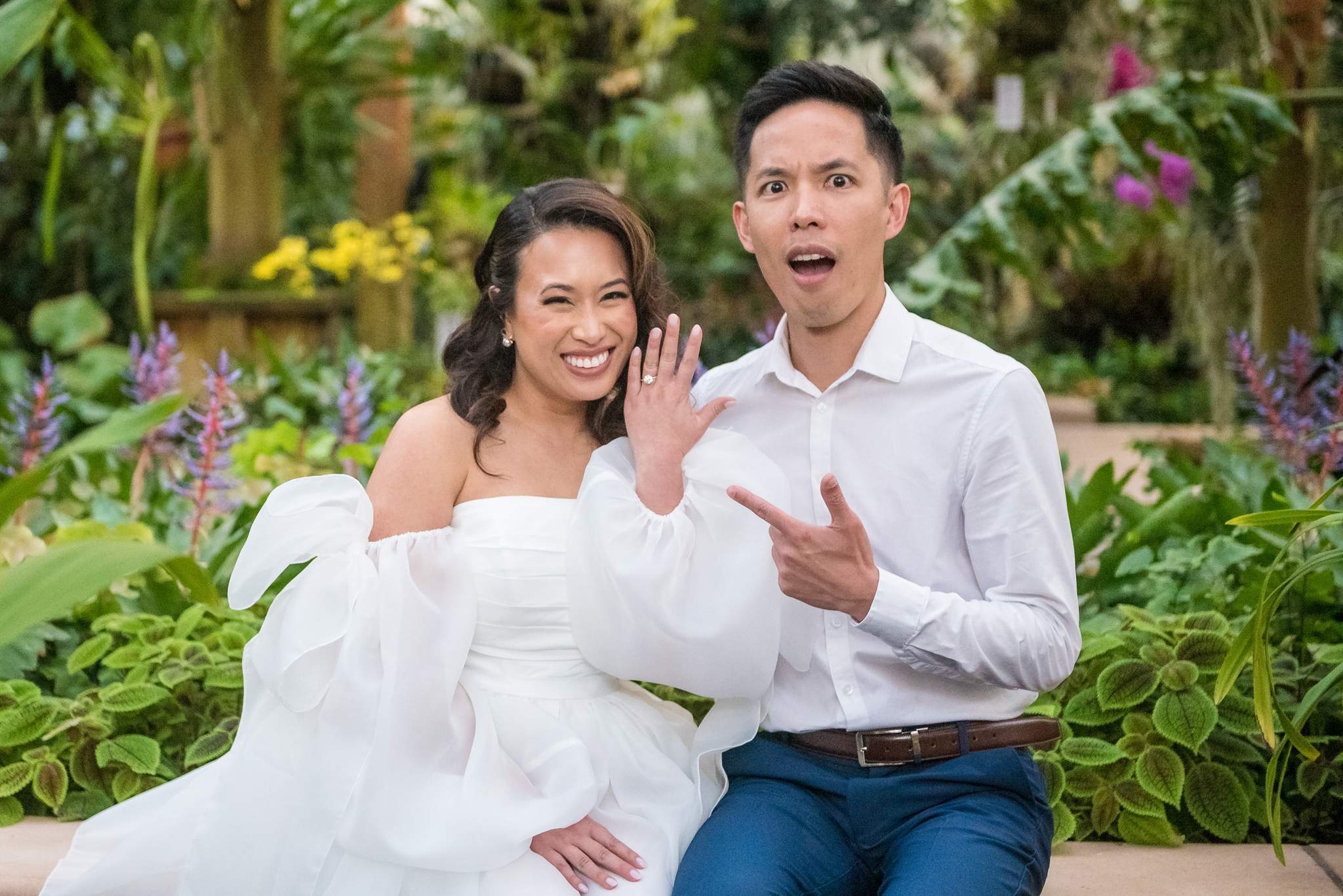 Bride-to-be showing off her ring with a surprised partner among tropical flowers