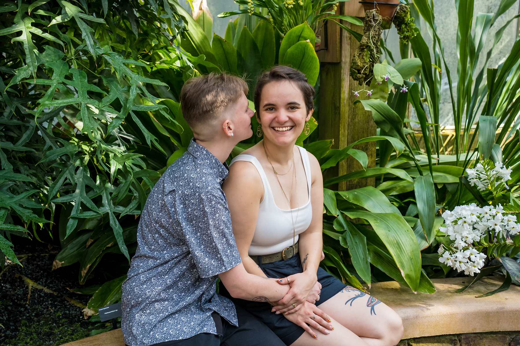 Engaged couple seated among tropical plants and white blooms in the Fuqua Conservatory