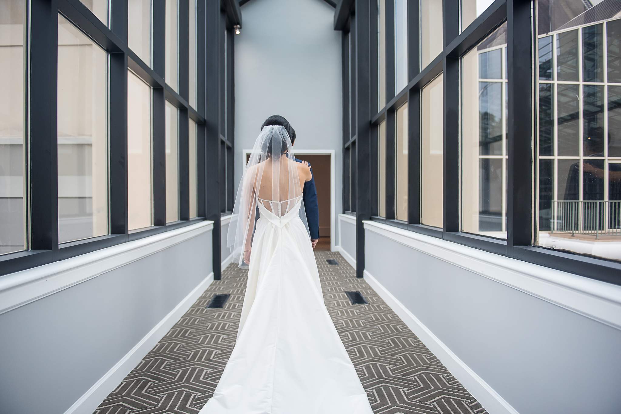 Bride in a backless gown walks down a glass and black-framed corridor at Chateau Elan