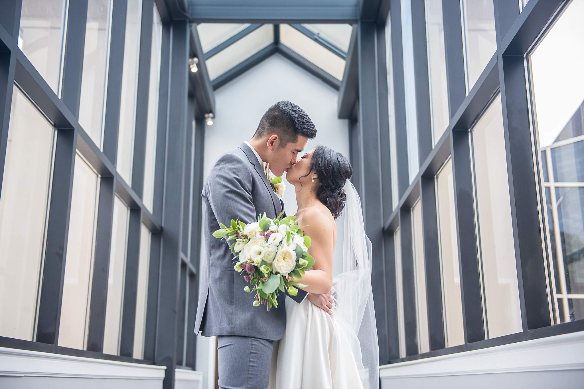 Bride and groom kiss beneath the peaked skylight of a glass corridor at Chateau Elan