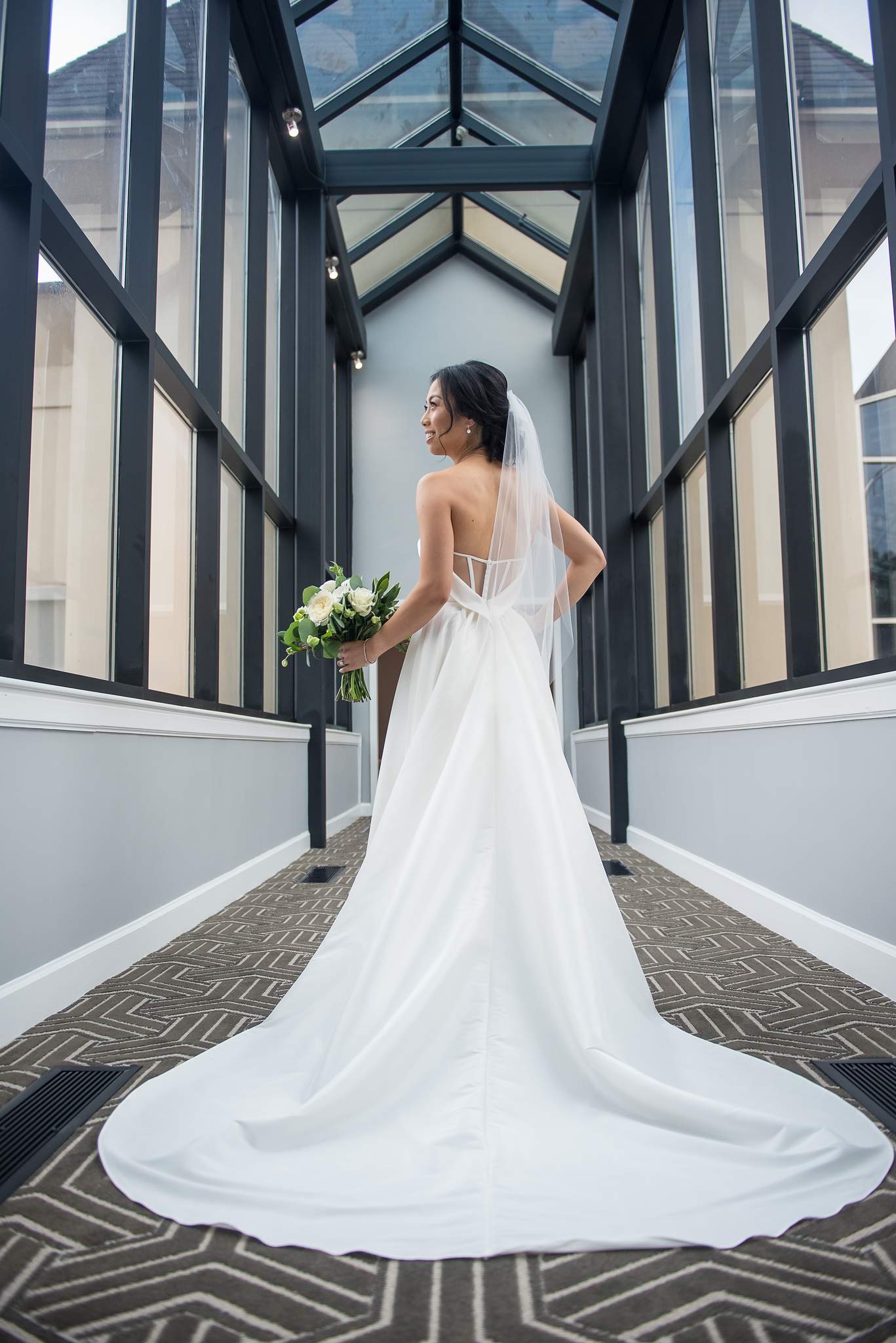 Bride's long satin train spreads across the patterned carpet of a glass corridor at Chateau Elan