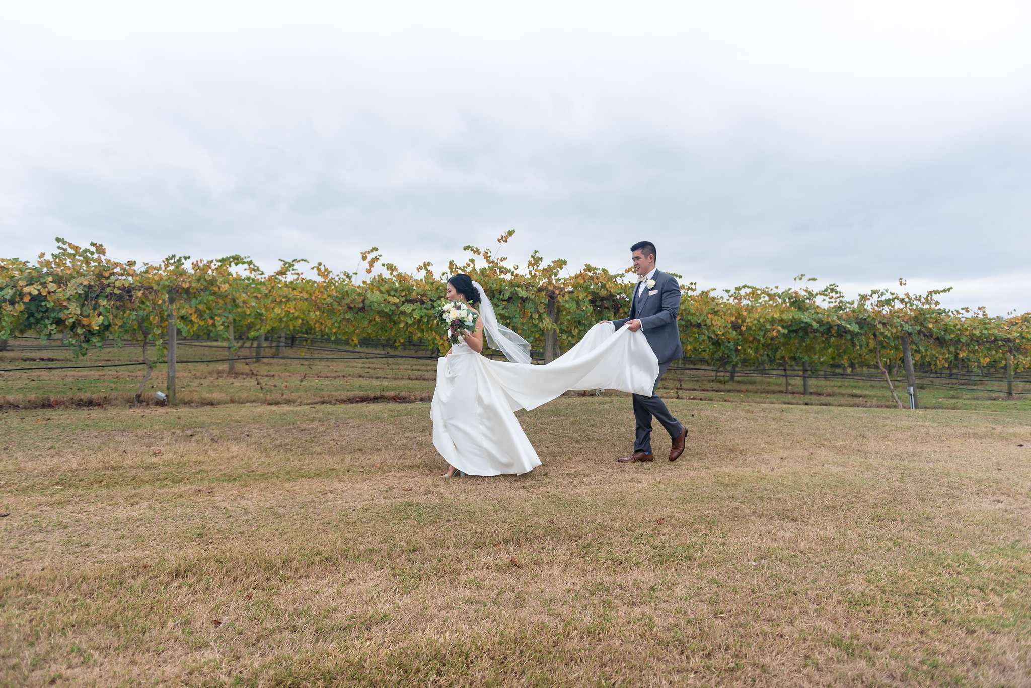 Groom carries the bride's flowing veil as they cross the field beside the Chateau Elan vineyard