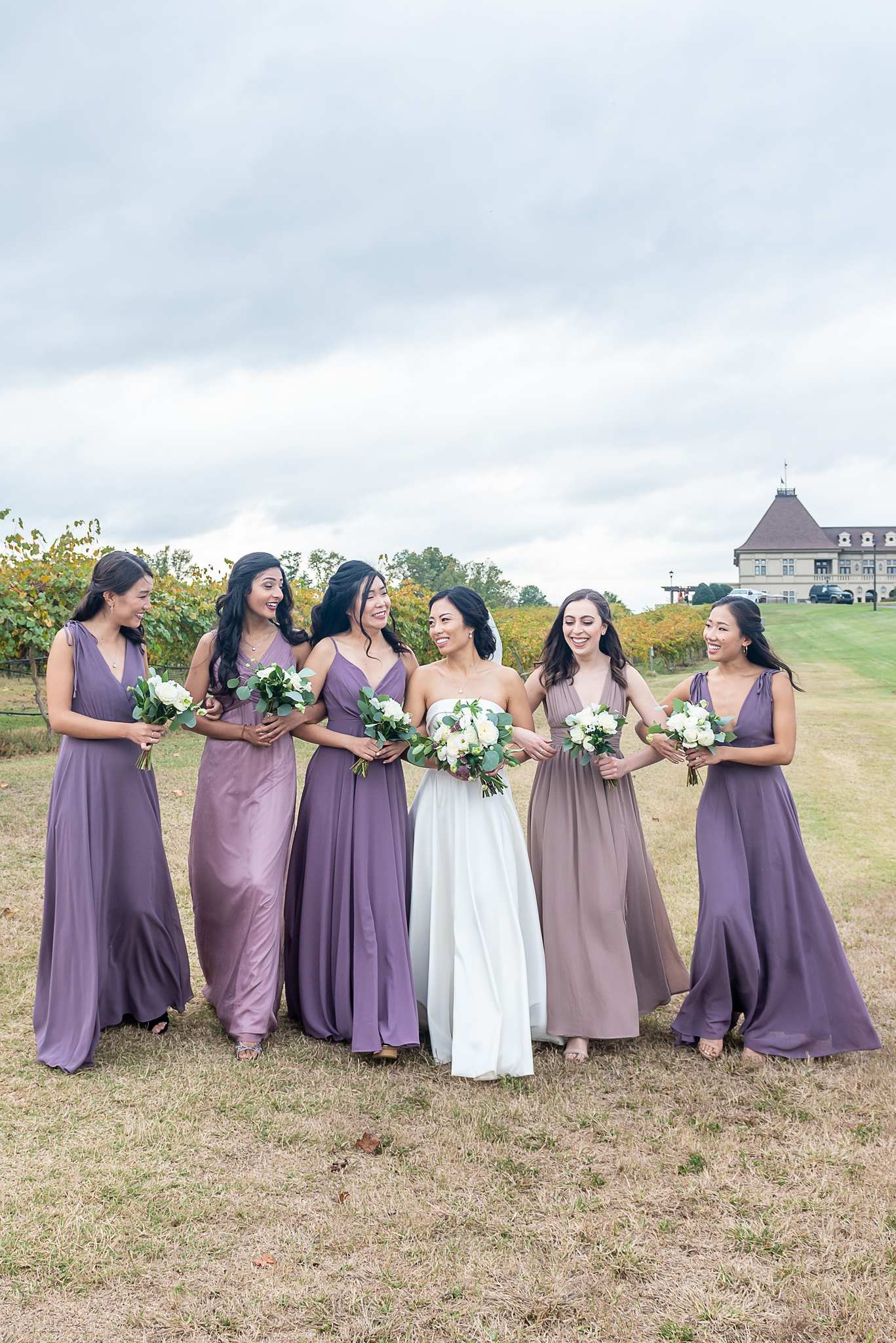 Bride walks with bridesmaids in lavender and mauve gowns through the vineyard toward Chateau Elan