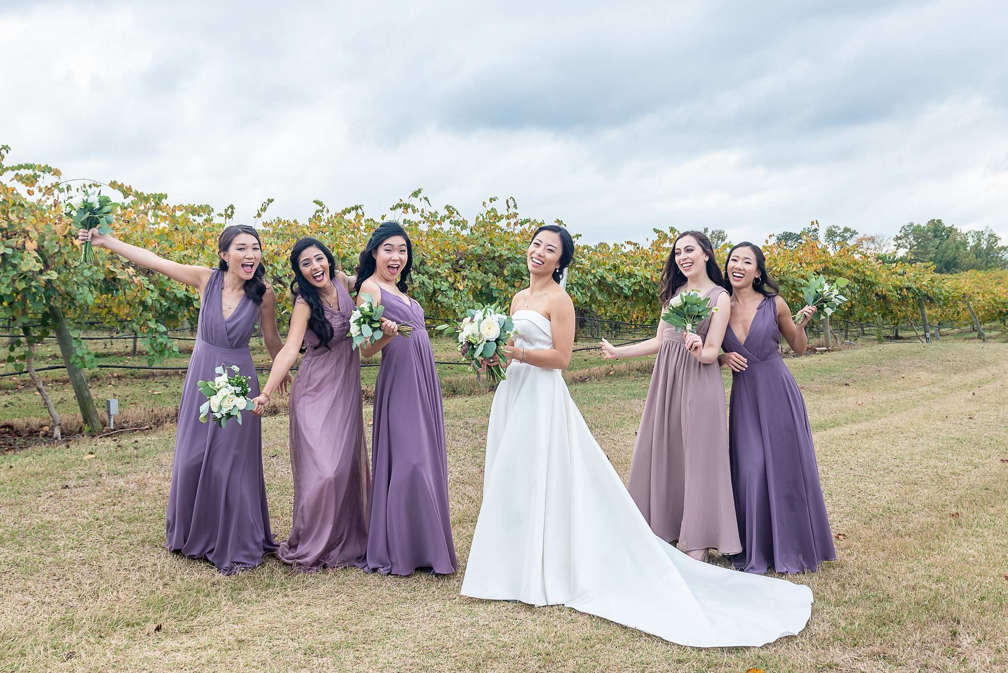 Bride laughs with bridesmaids in purple gowns among the vineyard rows at Chateau Elan
