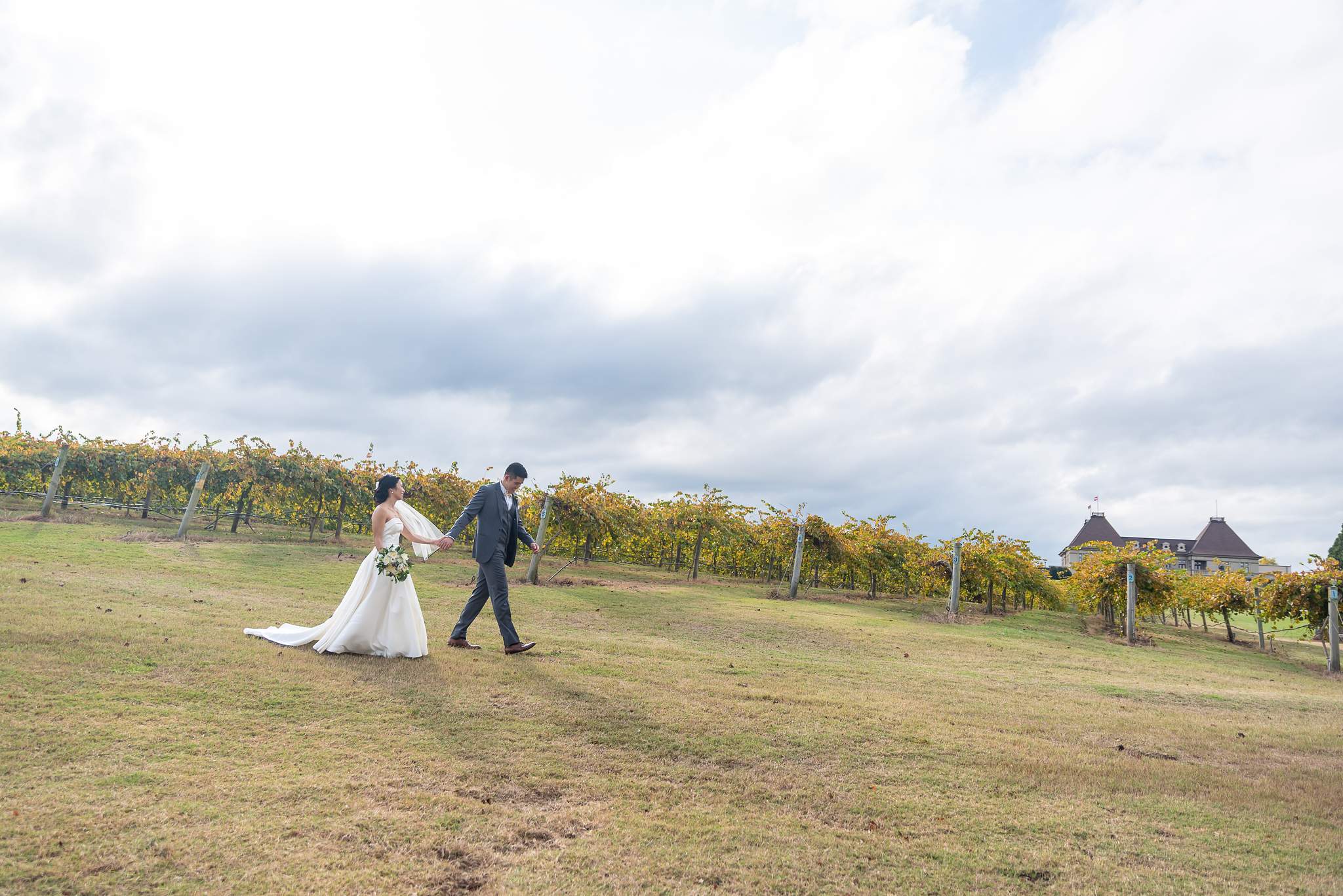 Bride and groom walk across the open vineyard field with Chateau Elan on the horizon