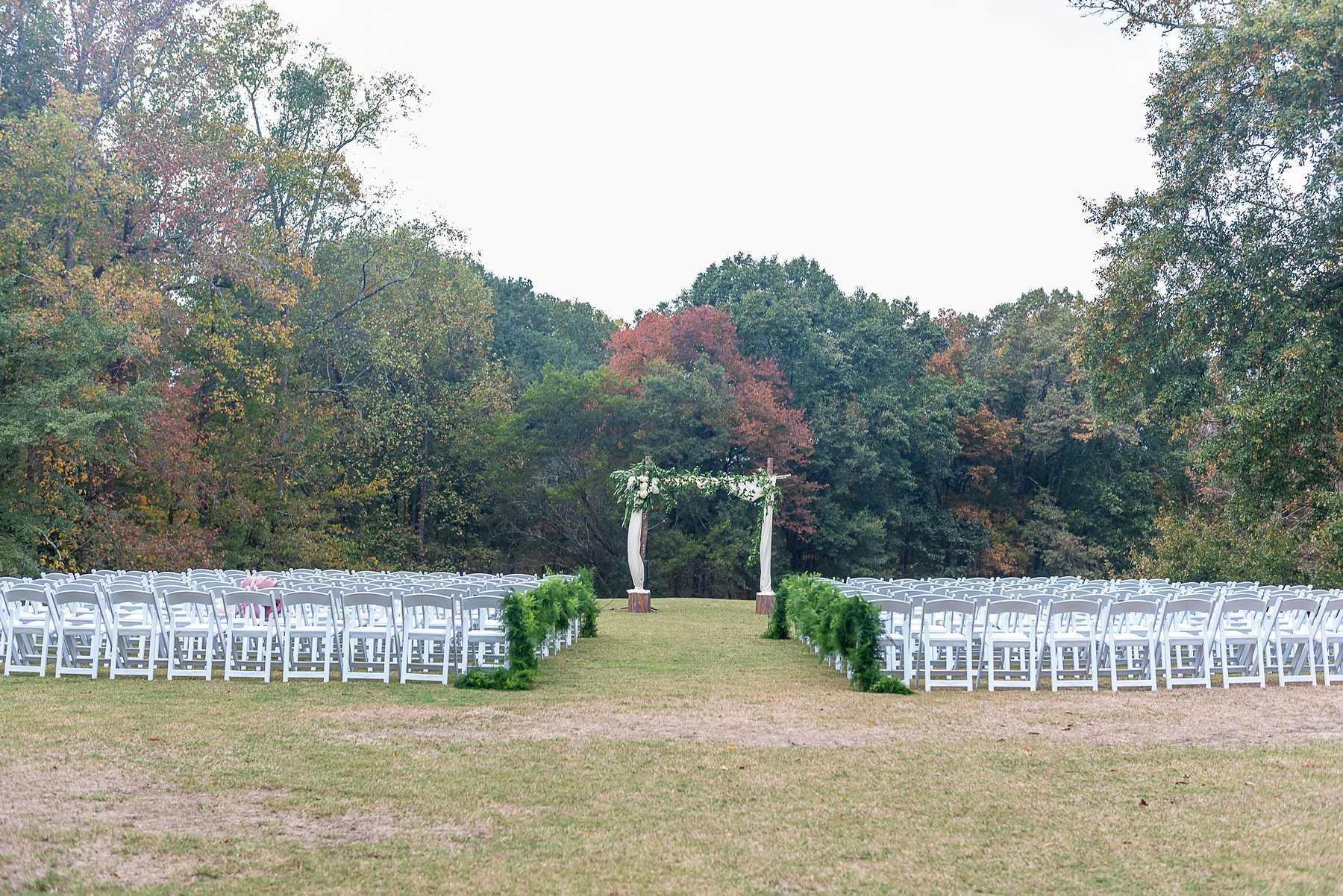 Empty outdoor ceremony site with white chairs, greenery and a floral arbor framed by autumn trees