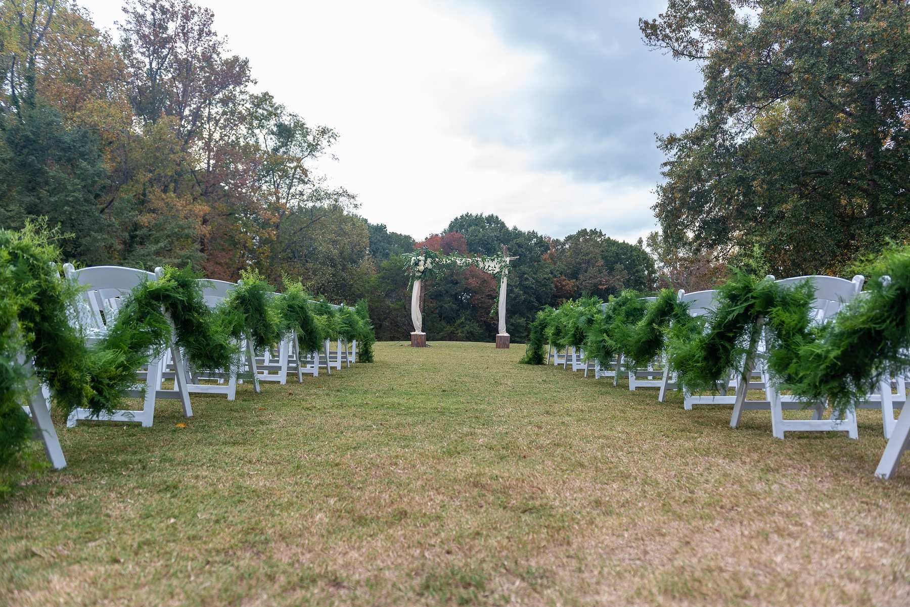 Ceremony aisle lined with white chairs draped in garland greenery leading to the floral arbor