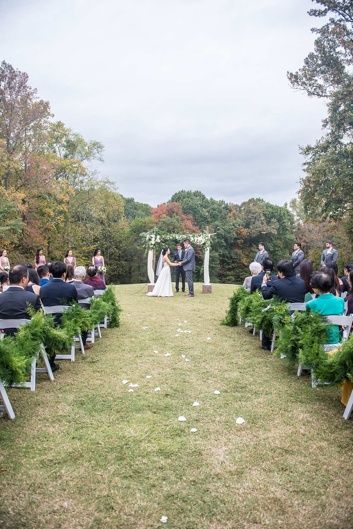 Vertical view down the petal-strewn aisle to the couple at the arbor during the outdoor ceremony