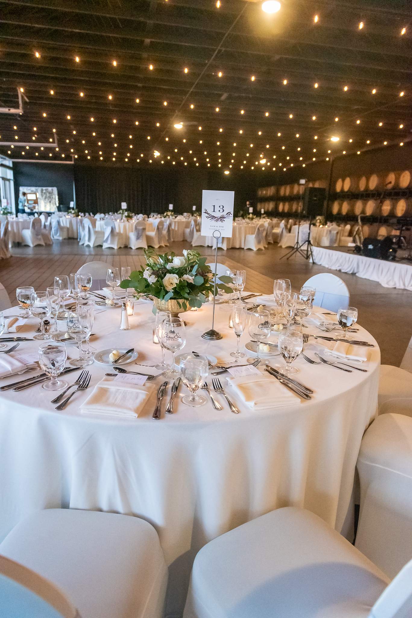 Reception table with glassware and white chair covers in a string-lit room lined with wine barrels