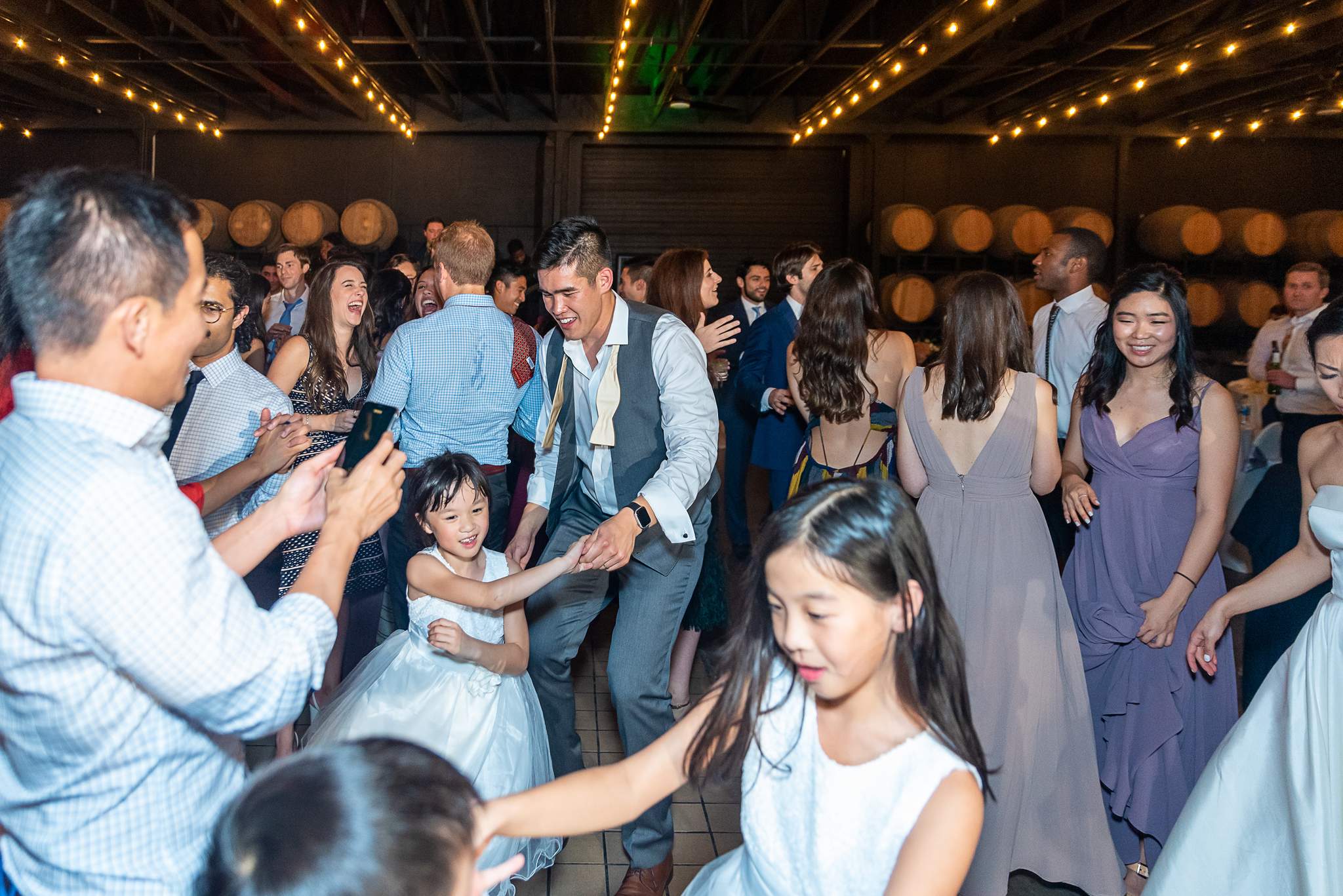 Guests and a young flower girl dance together under string lights near the wine barrels