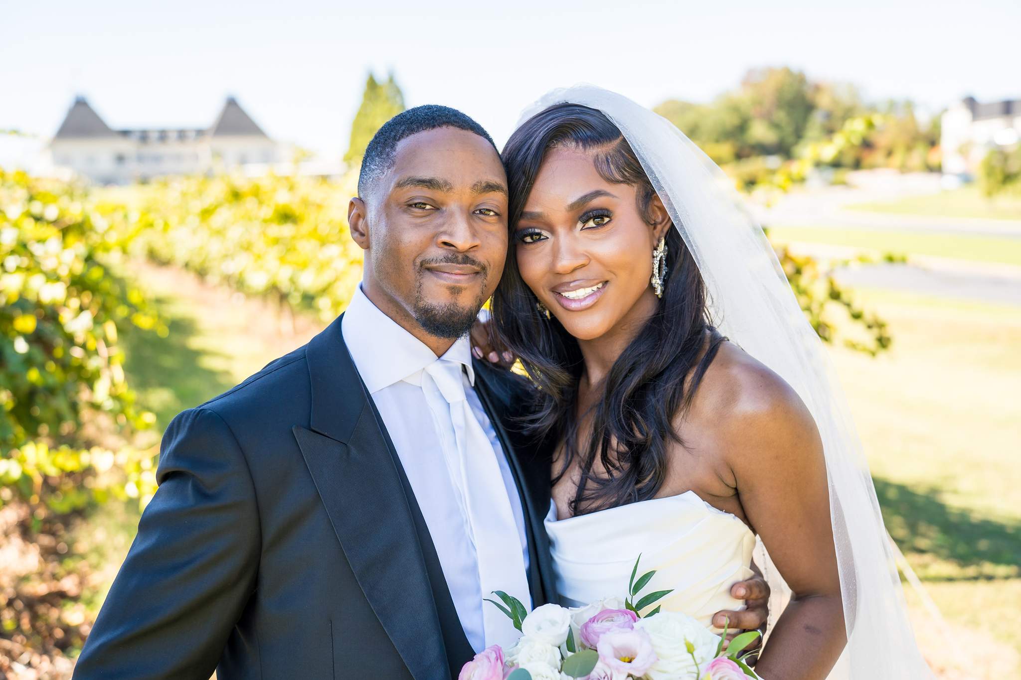 Smiling bride and groom pose cheek to cheek with the vineyard and chateau behind them