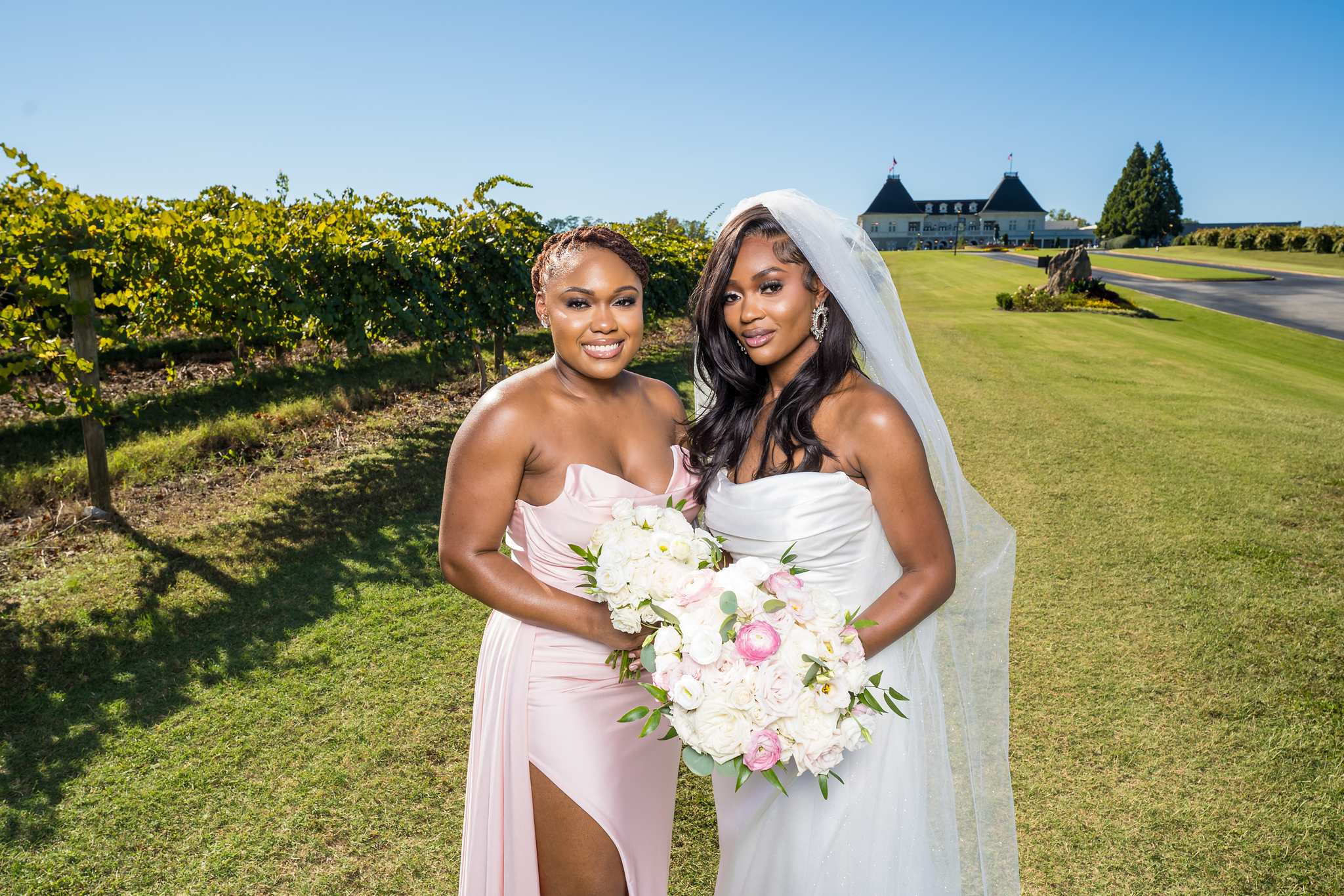Bride and a bridesmaid in a blush gown smile together in the vineyard on a sunny day