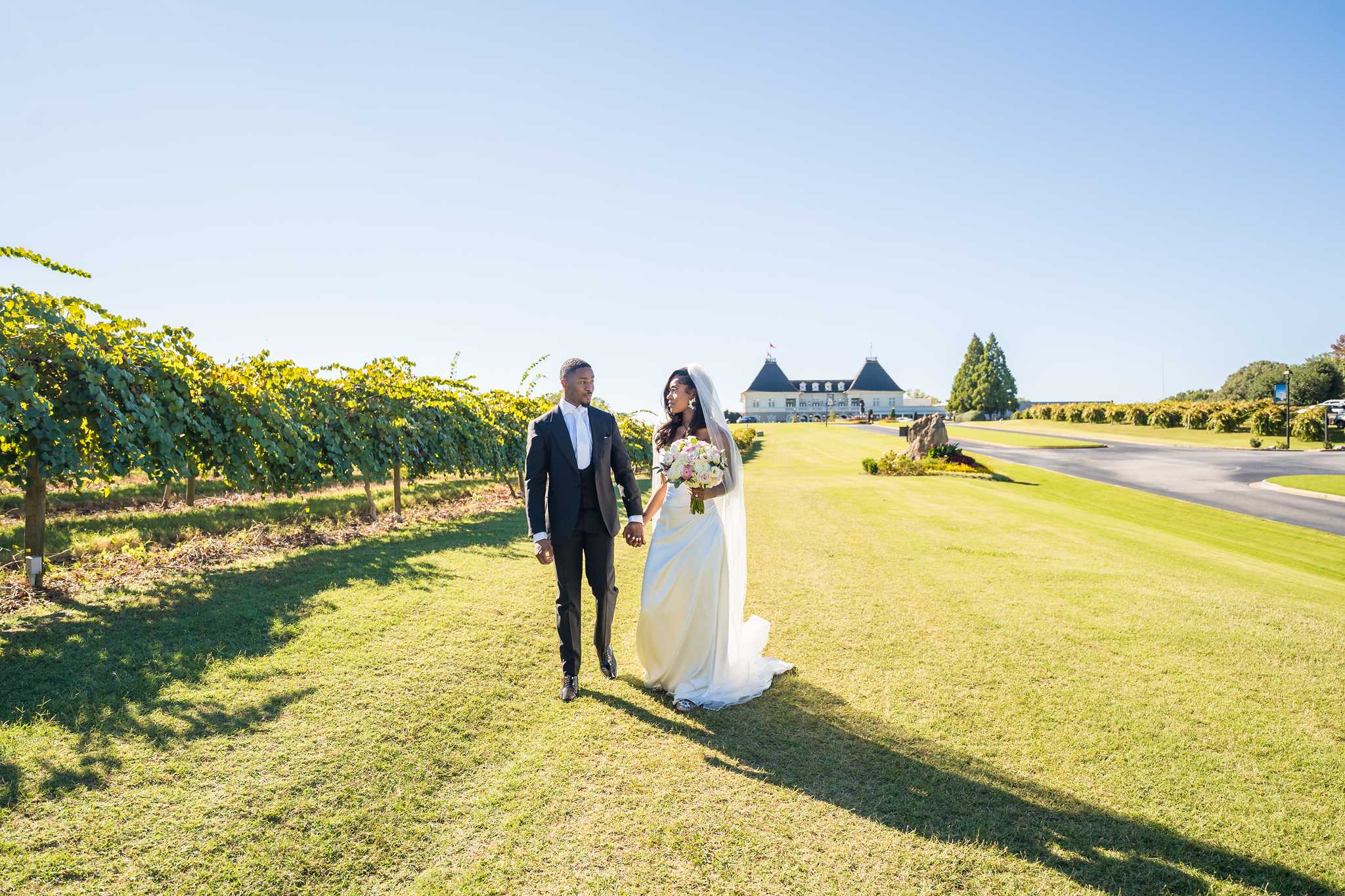 Bride and groom walk hand in hand across the sunny vineyard lawn with Chateau Elan behind them
