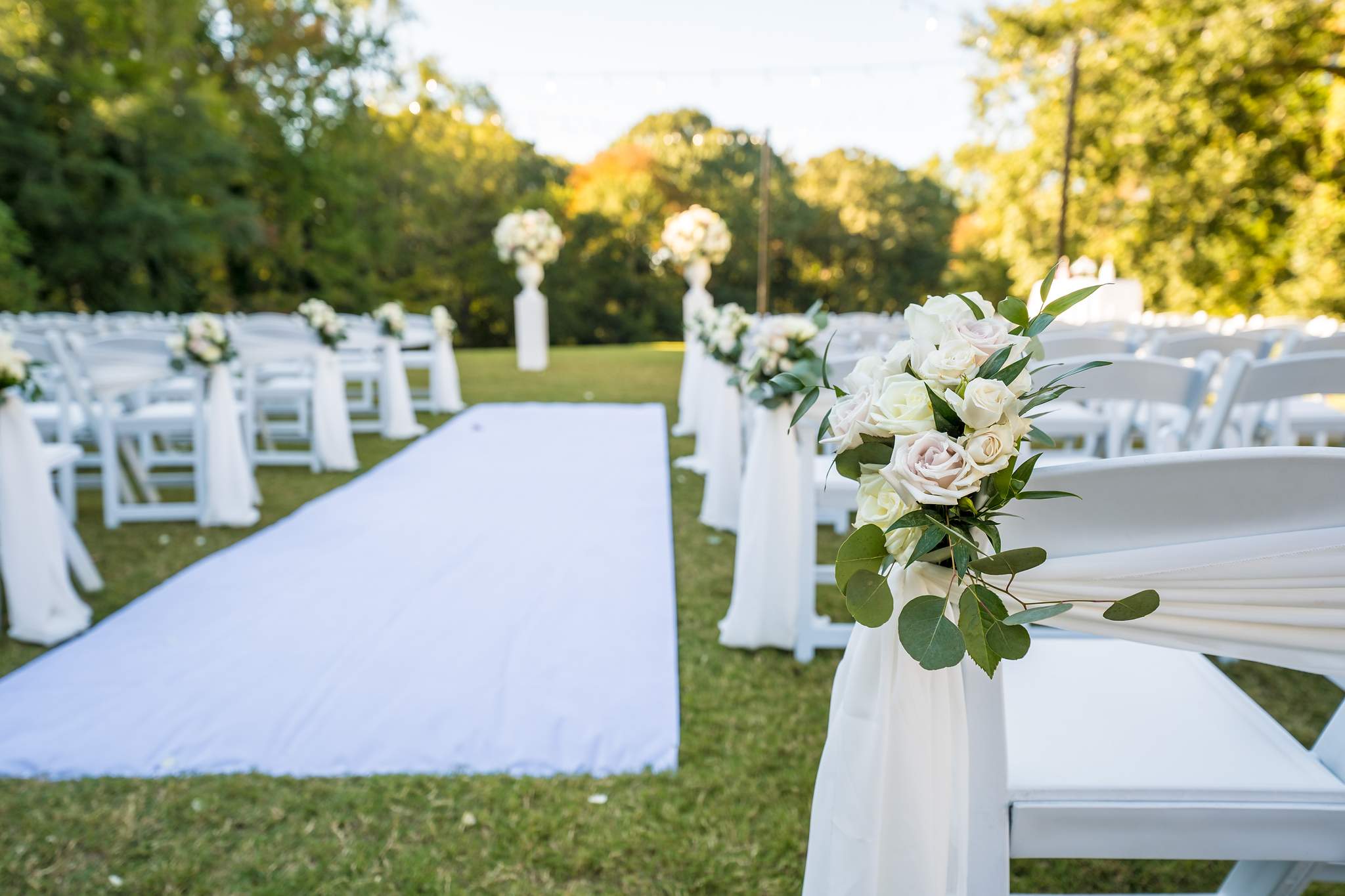 White aisle runner lined with white chairs, draped fabric and white rose arrangements on the ceremony lawn