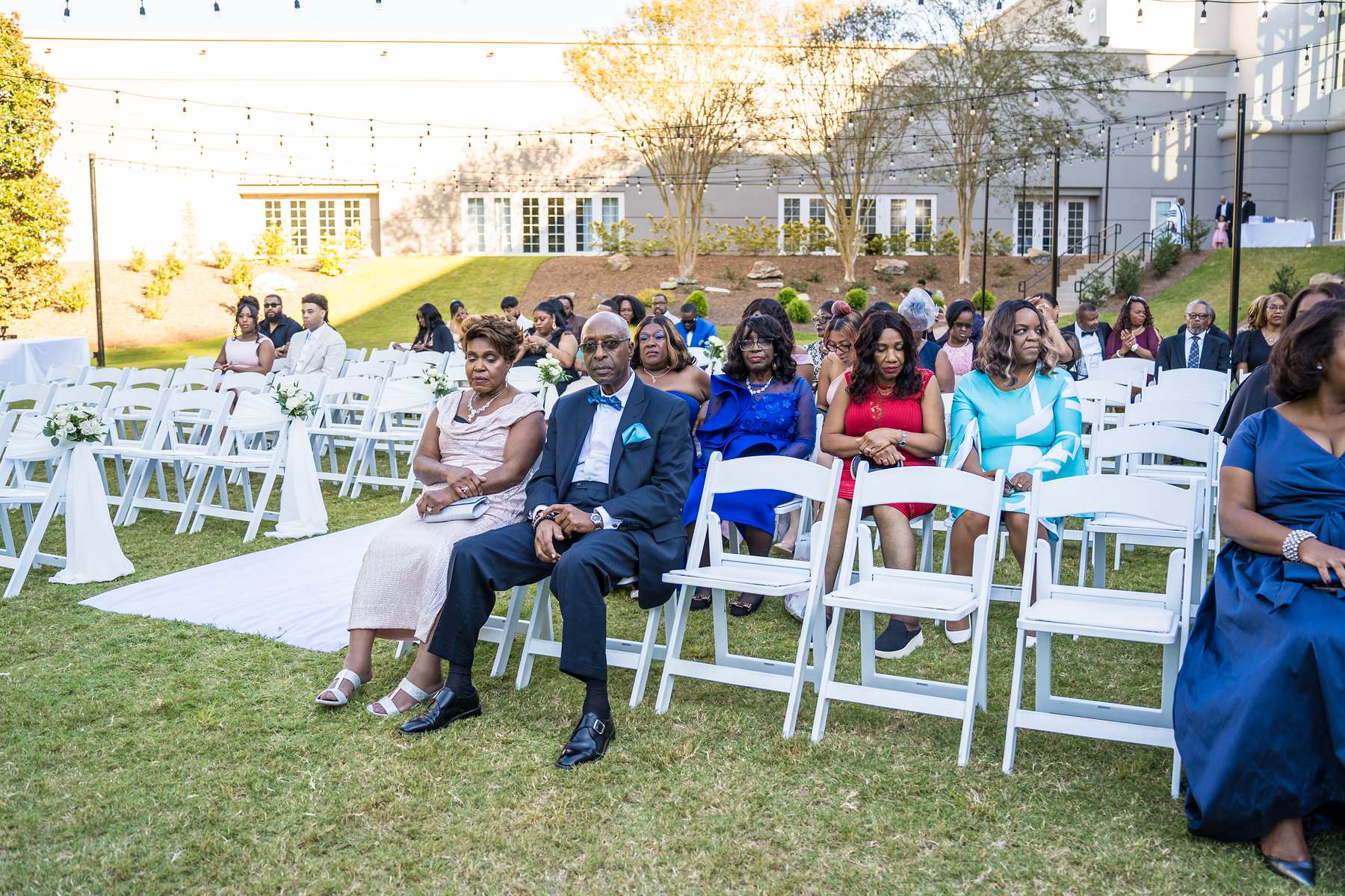 Guests seated in white chairs on the ceremony lawn wait for the wedding to begin