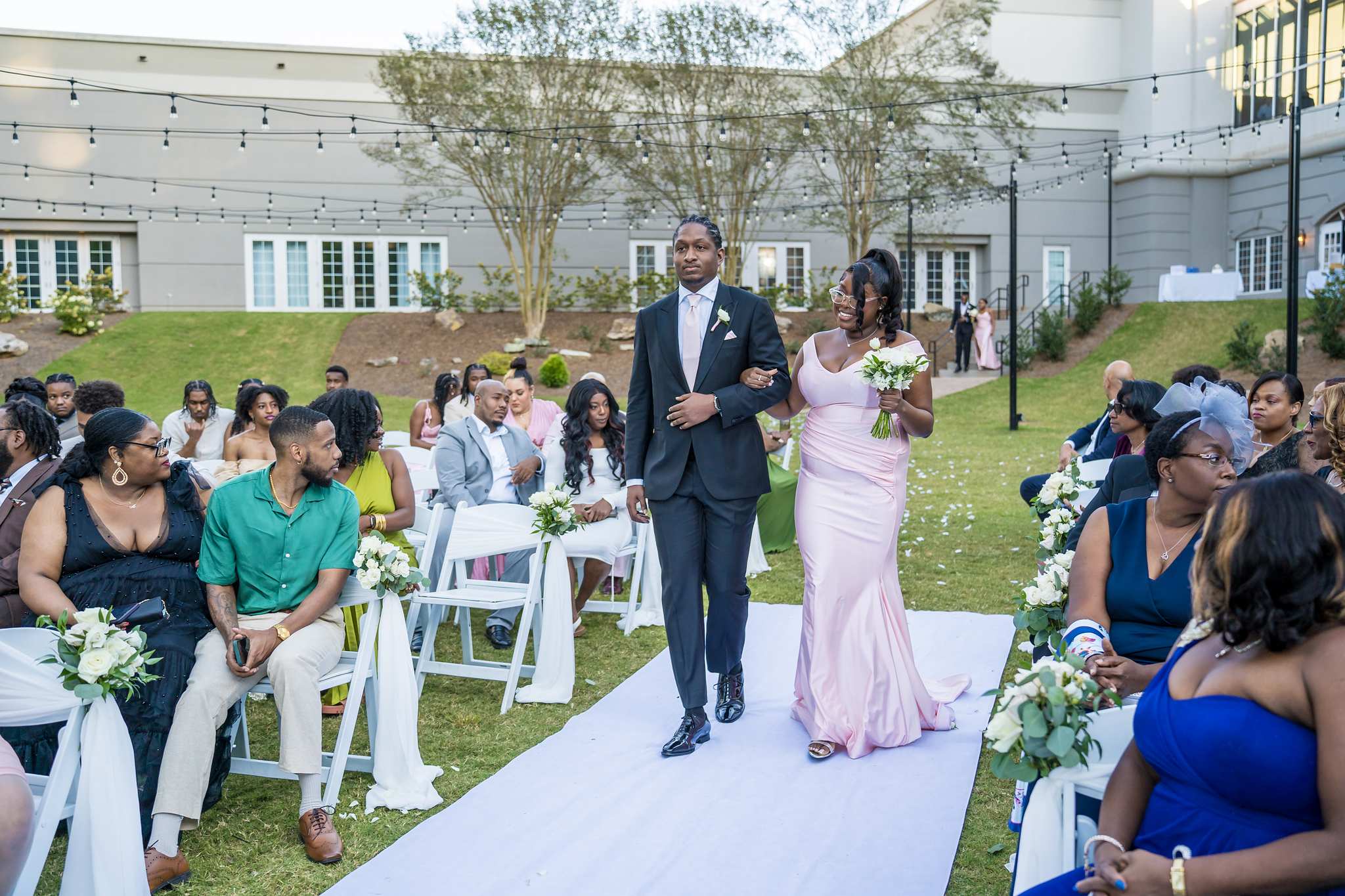 Groomsman and bridesmaid in blush pink walk down the white aisle runner during the processional
