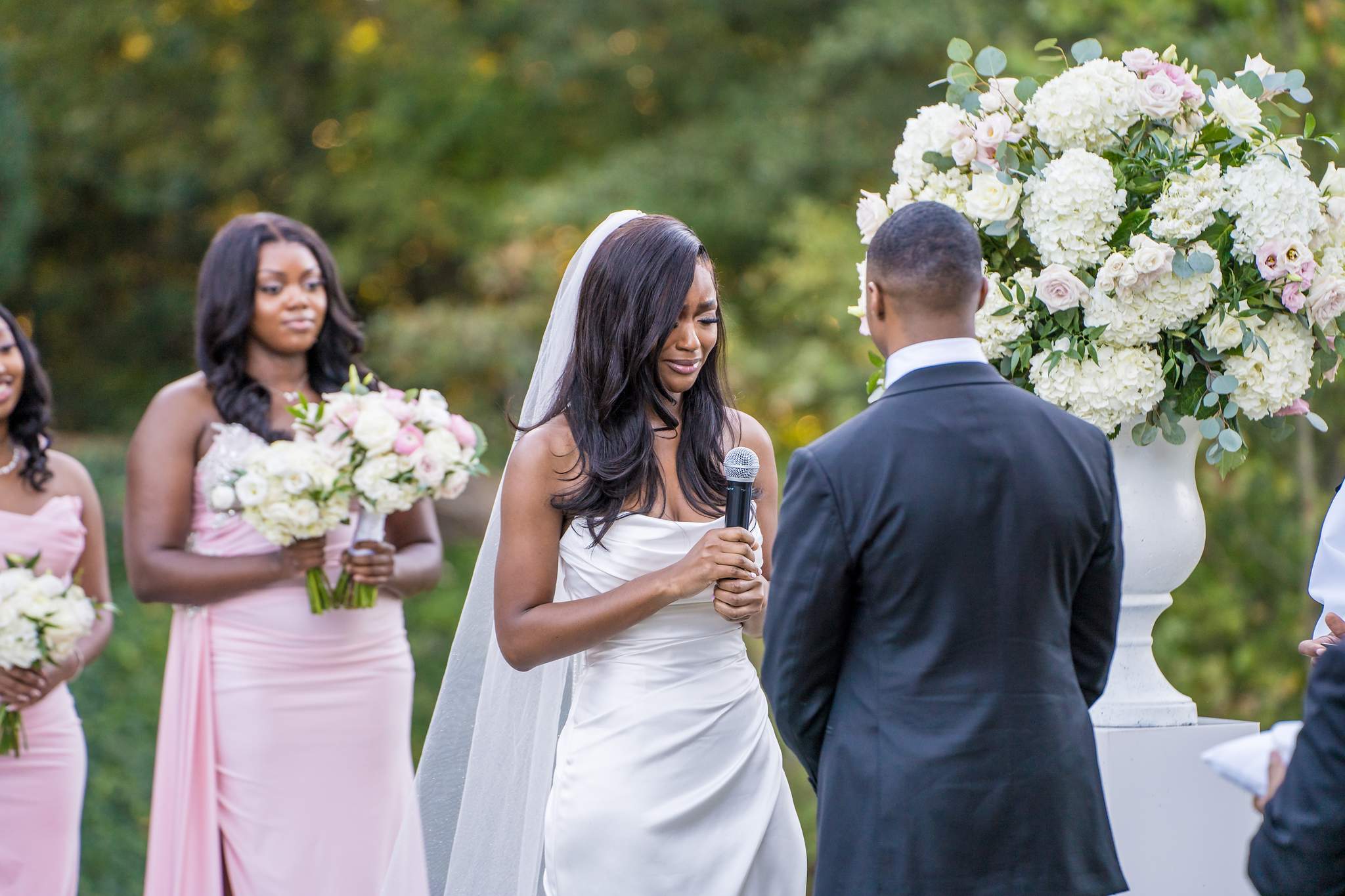 Bride reads her vows into a microphone as the groom and a bridesmaid look on