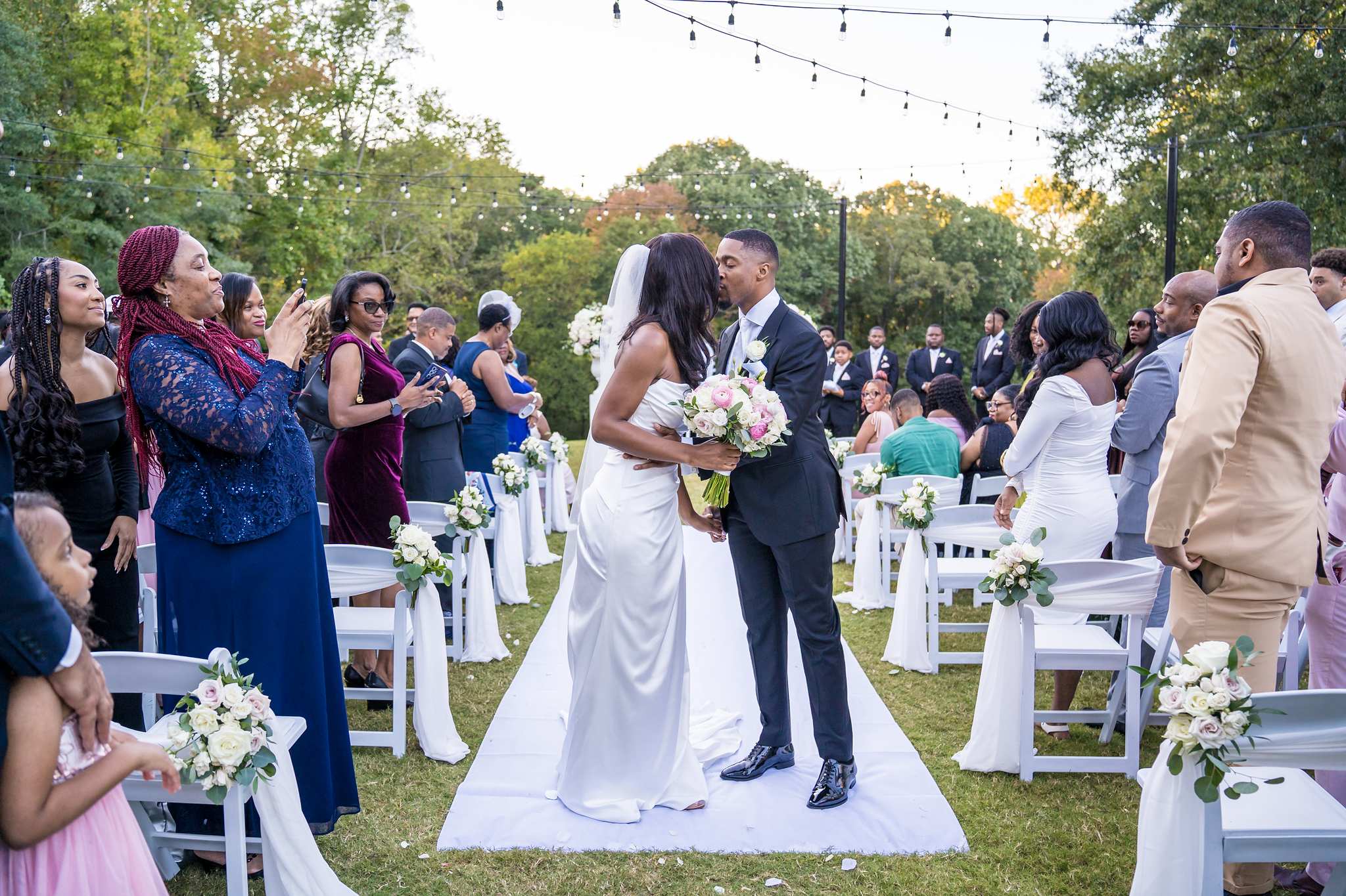 Bride and groom kiss on the white aisle runner as guests cheer and take photos