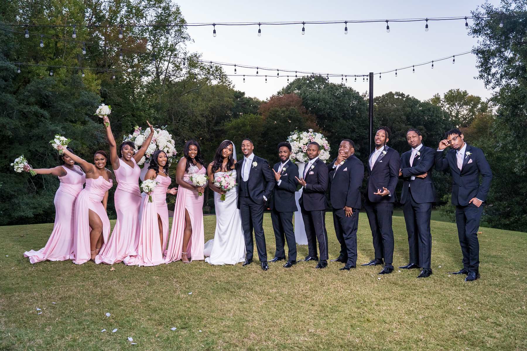 Bridesmaids in blush pink and groomsmen in black tuxedos cheer on the lawn under string lights