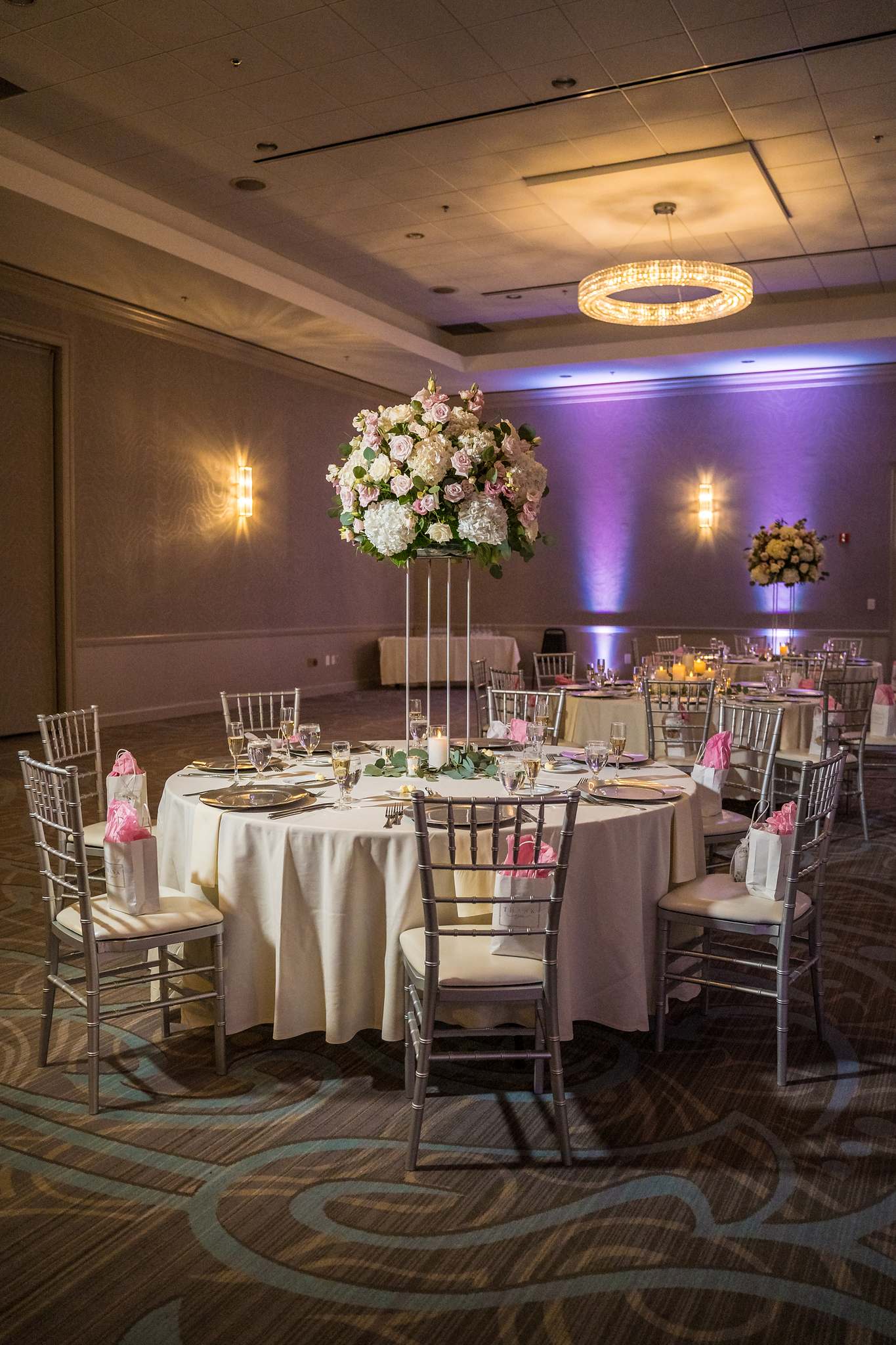 Round reception table with a tall pink and white centerpiece under purple uplighting