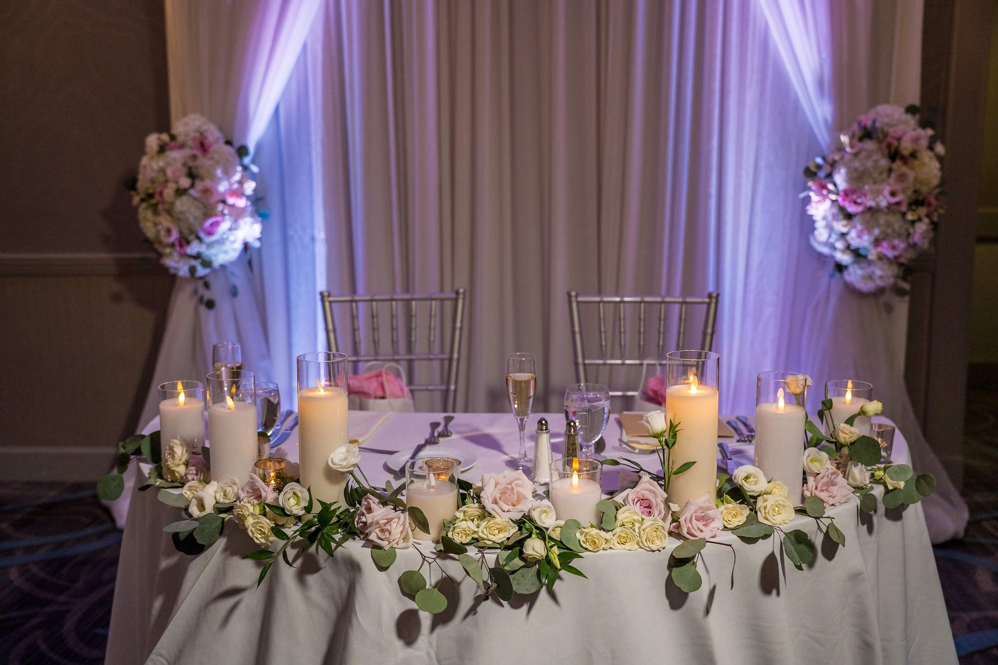 Sweetheart table with pillar candles, roses and greenery in front of white draped fabric