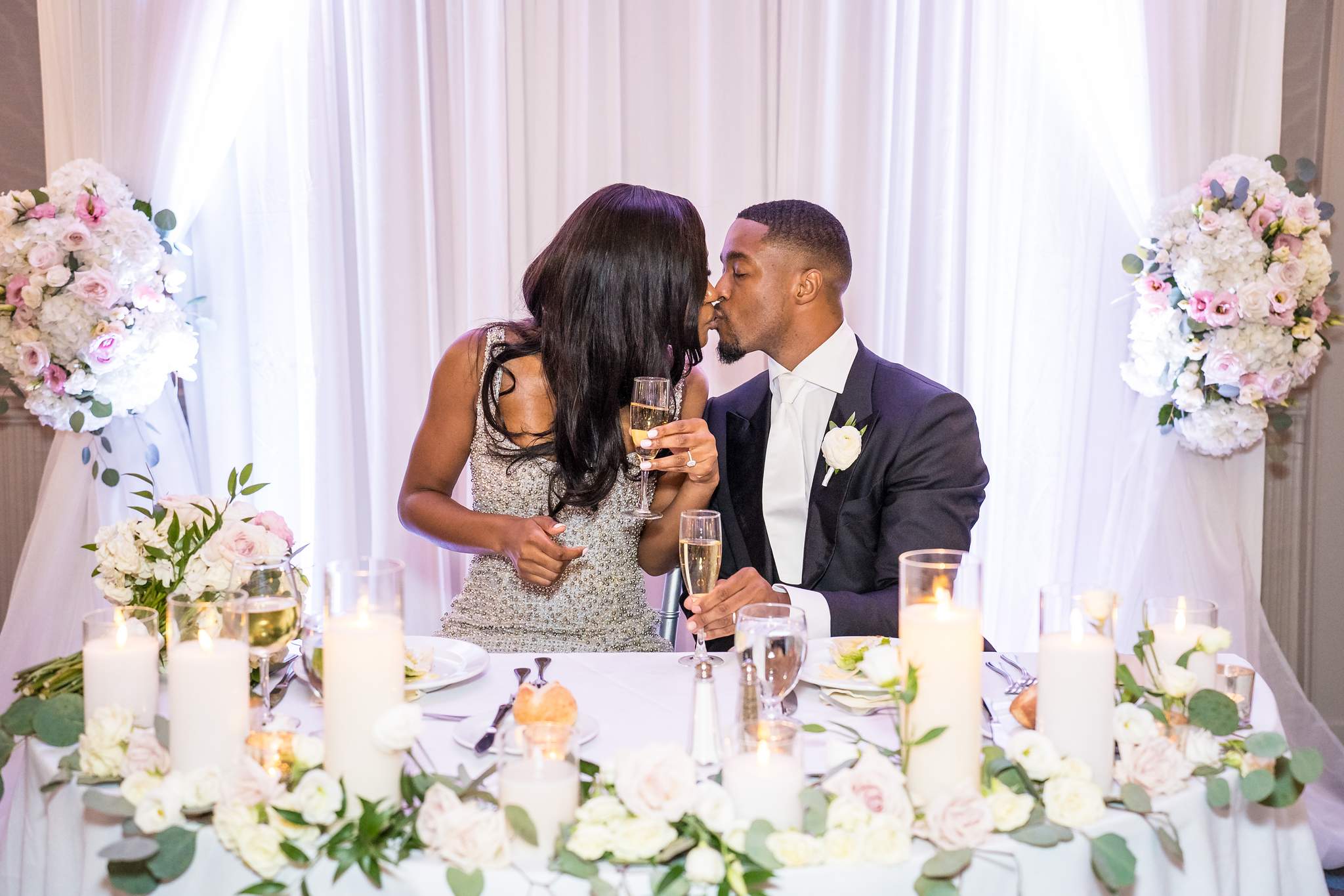 Bride and groom kiss at the candlelit sweetheart table surrounded by white and pink flowers