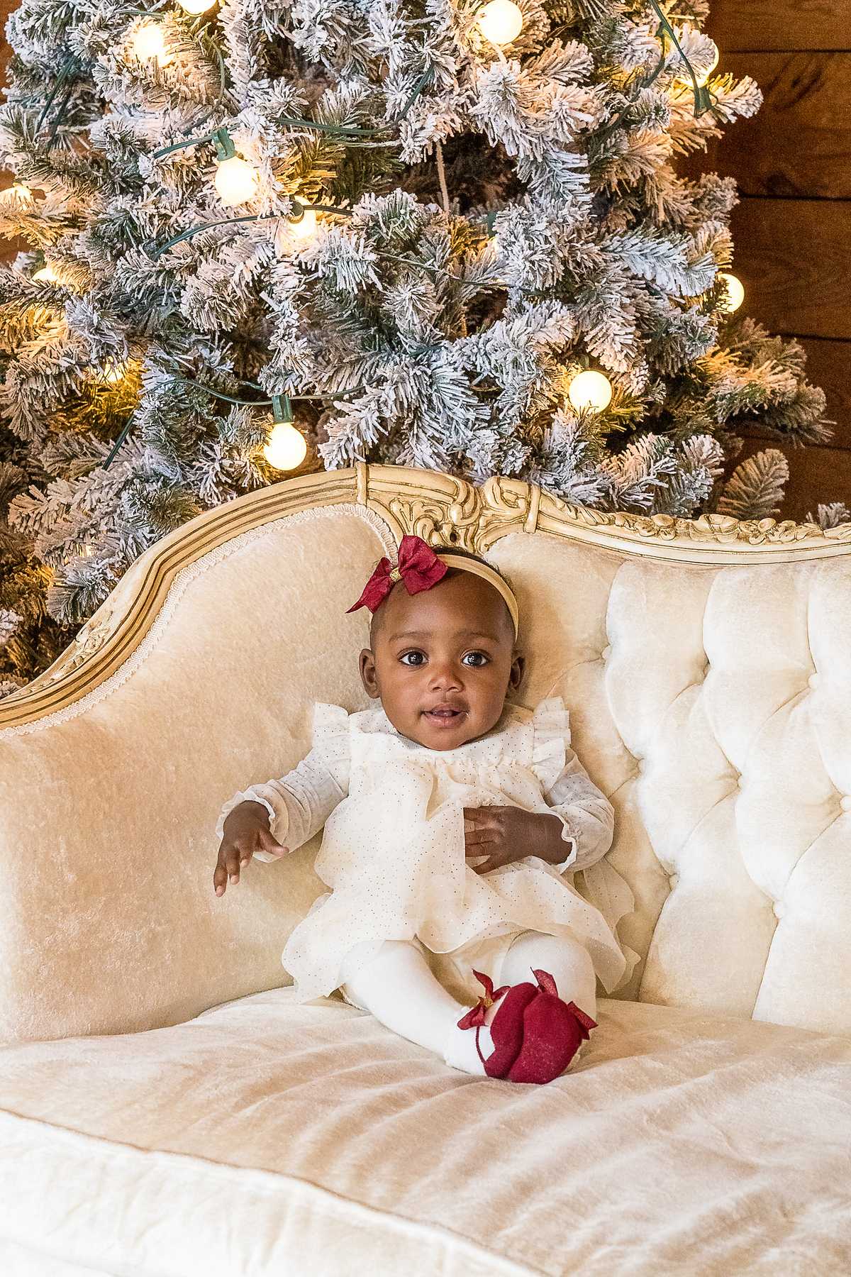 Baby in an ivory outfit and red shoes on a cream tufted settee in the Christmas Barn