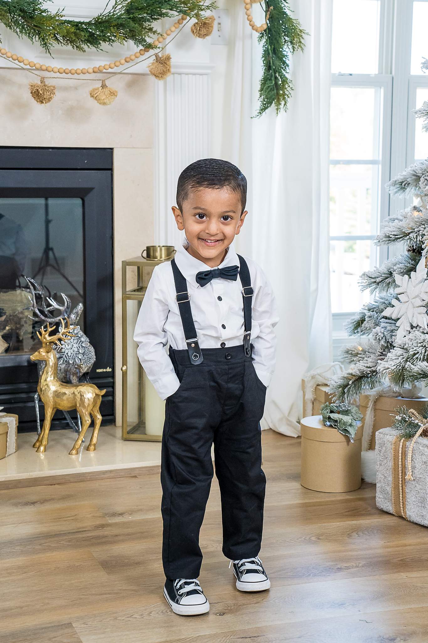 Little boy in a white shirt, black bow tie and suspenders beside a Christmas tree