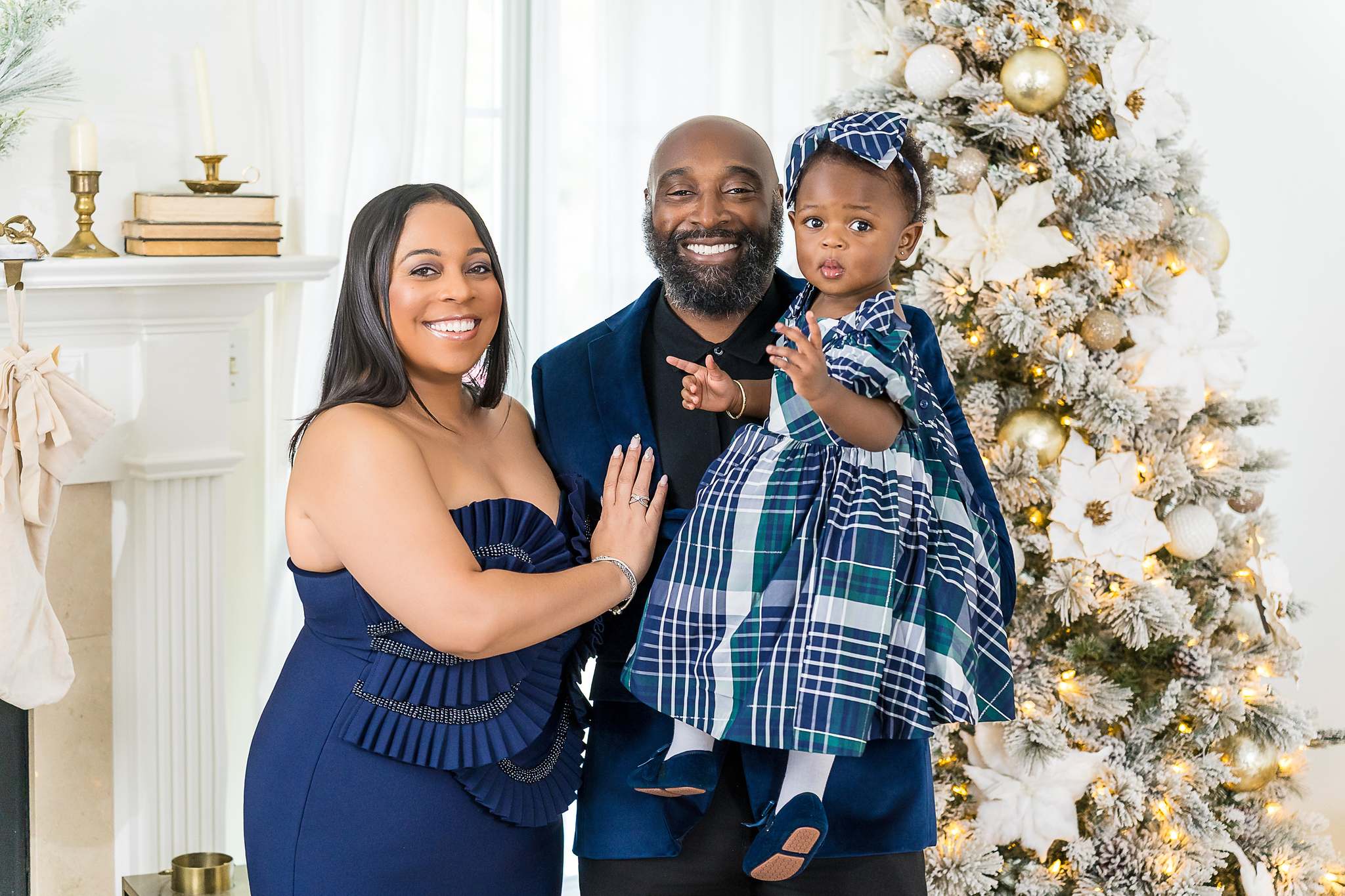 Parents in navy velvet with their toddler in a blue plaid dress beside a Christmas tree