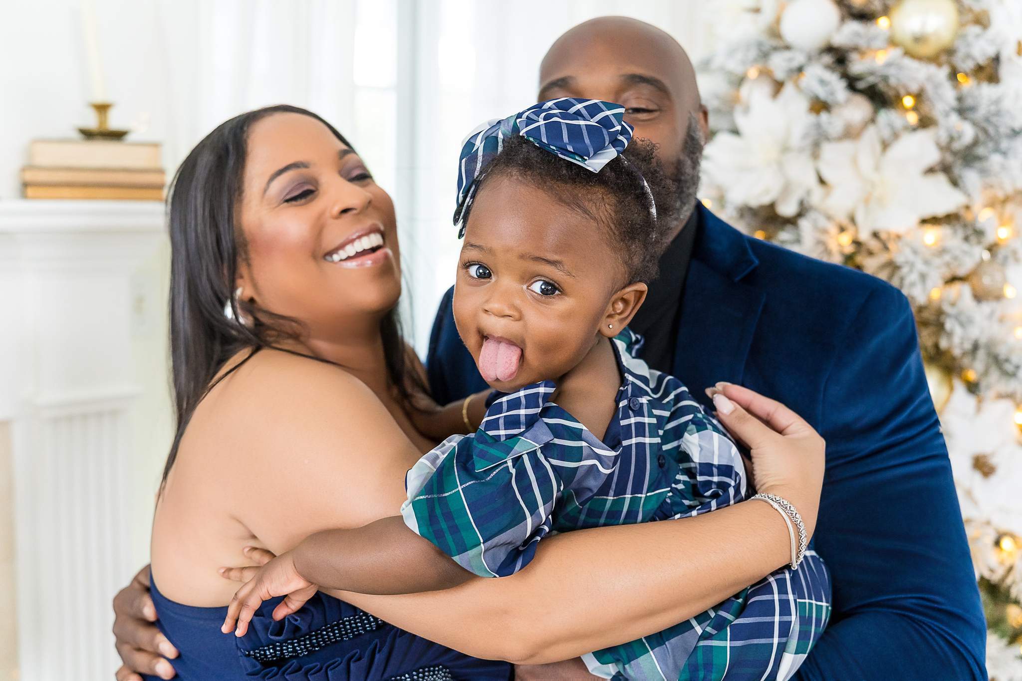 Parents in navy velvet hugging their laughing toddler in front of a flocked Christmas tree with gold ornaments