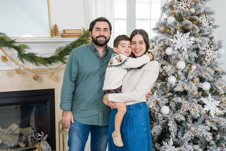 Family of three beside a flocked Christmas tree and fireplace