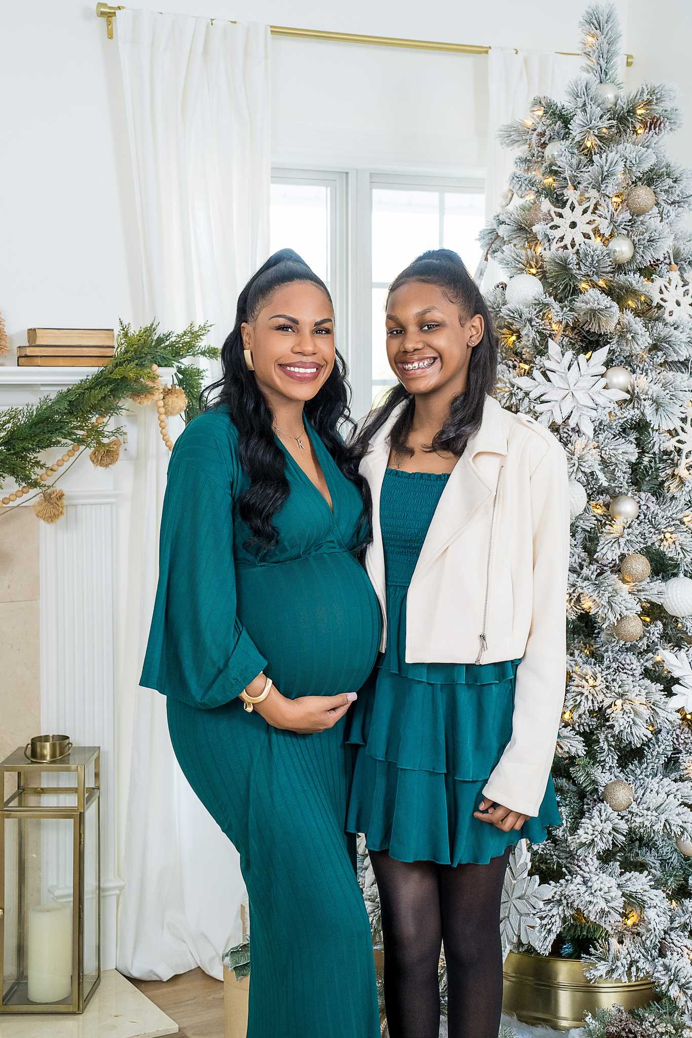 Expecting mother in a teal gown with her sister in teal and cream beside a Christmas tree