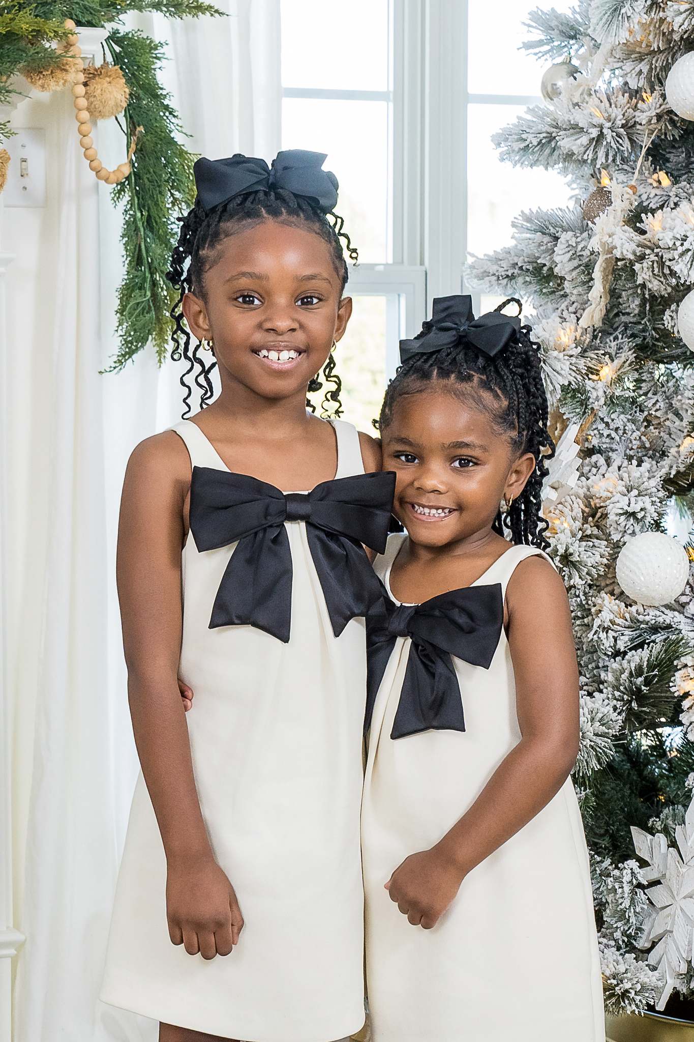 Two sisters in white dresses with oversized black bows beside a Christmas tree