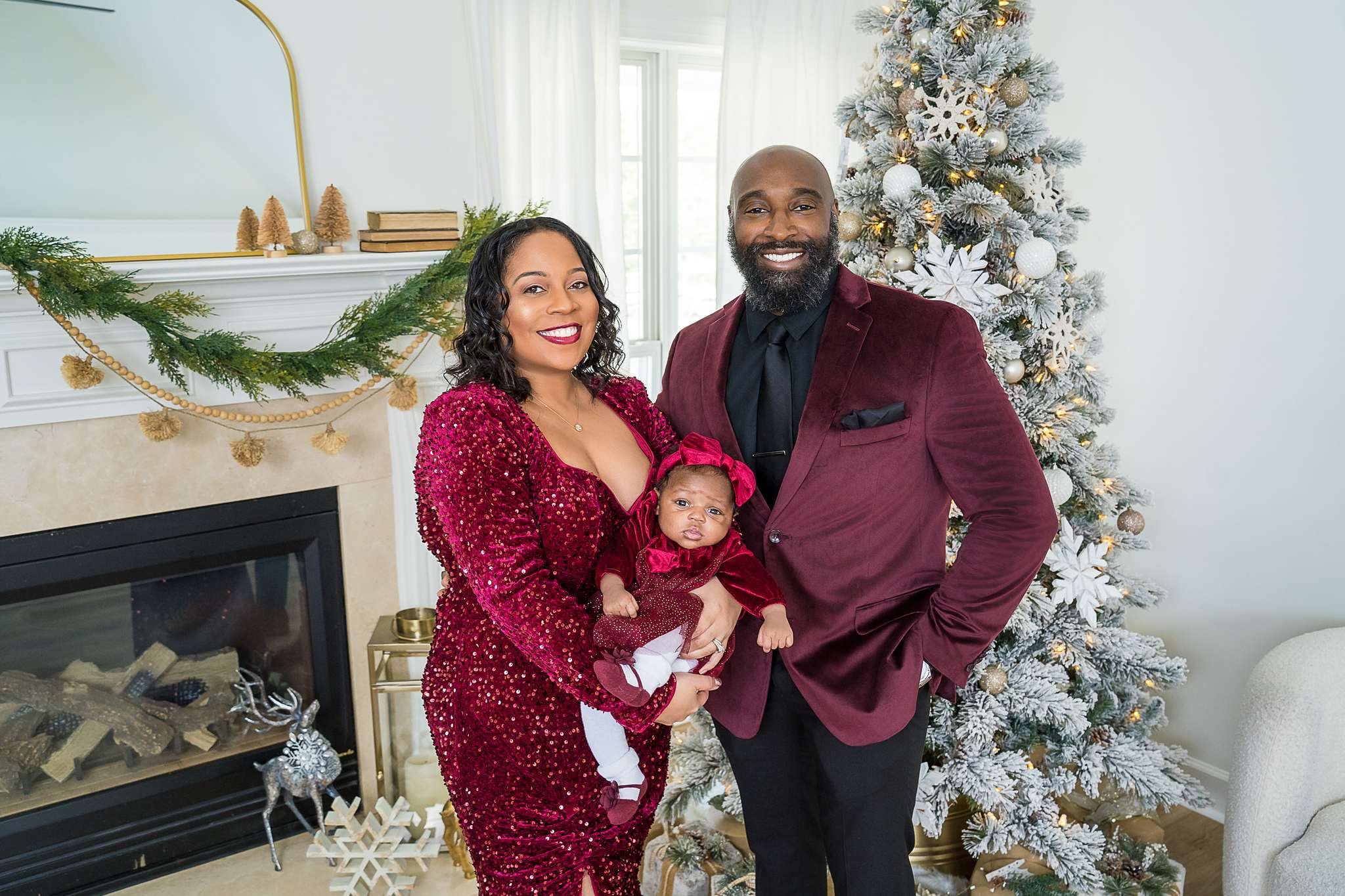 Couple in coordinating burgundy velvet holding their baby by a Christmas tree