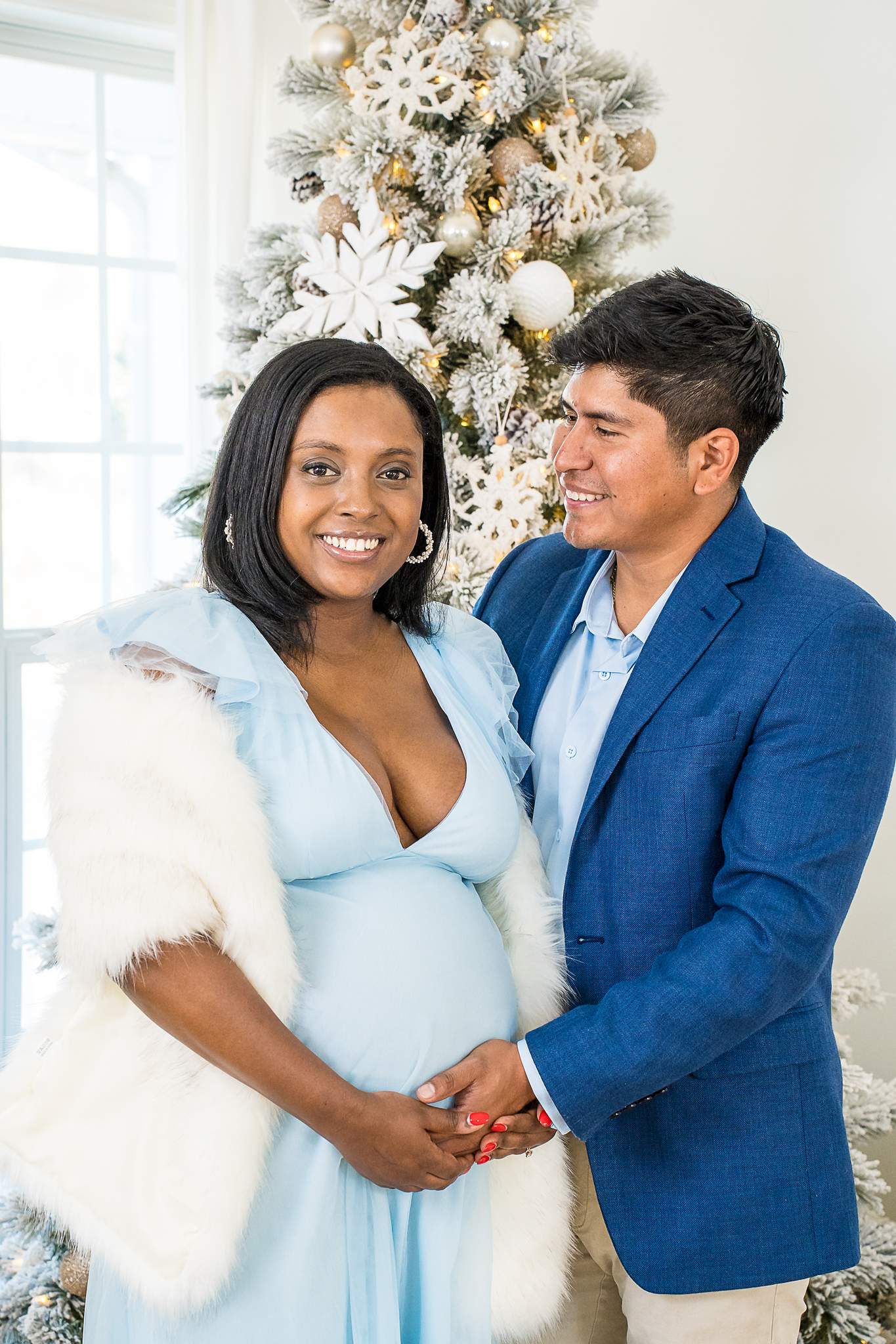 Expecting couple in icy blue and navy in front of a white Christmas tree