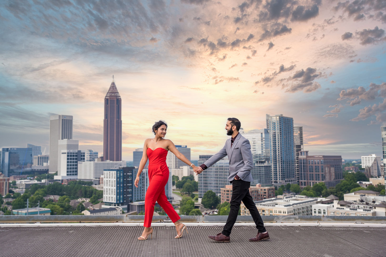 Engaged couple walking hand in hand across a rooftop with the Atlanta skyline and a dramatic sunset sky