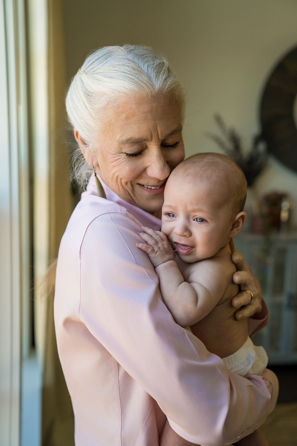 Grandmother in pink cradling her infant grandchild near a sunny window