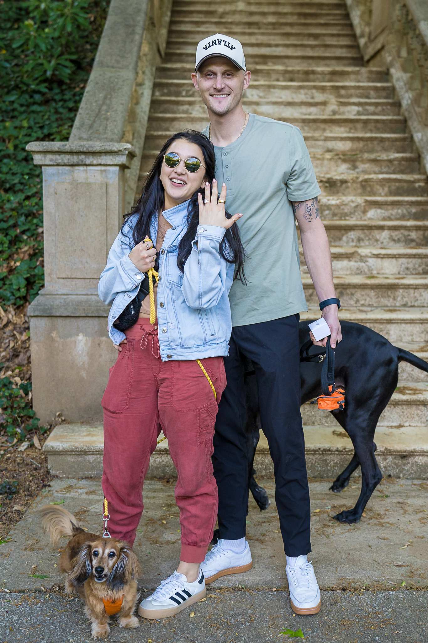 Smiling fiancee showing her ring beside her partner and their two dogs at the stone staircase at Cator Woolford Gardens