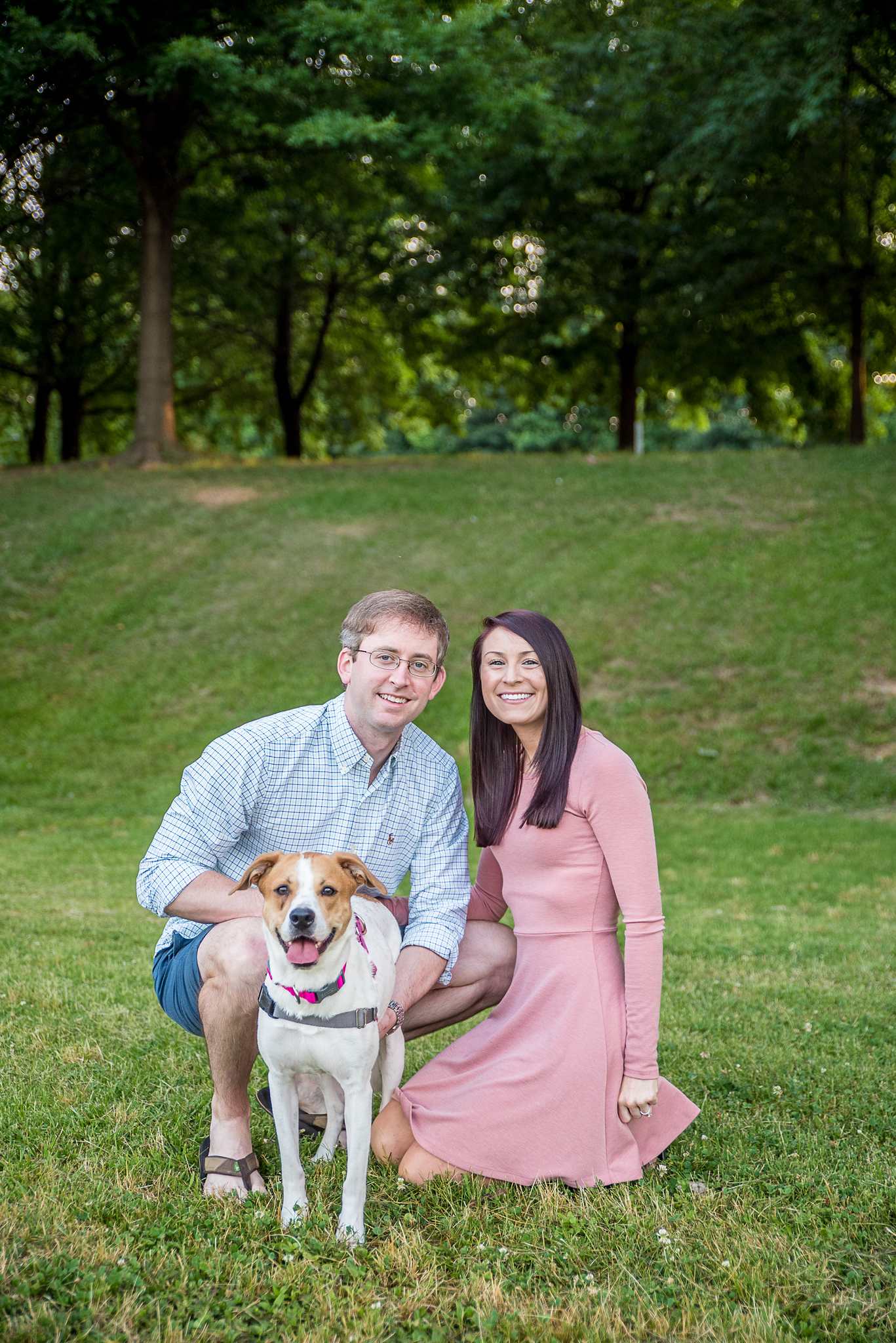 Couple crouching beside their smiling hound mix on a grassy hillside at Piedmont Park in Atlanta