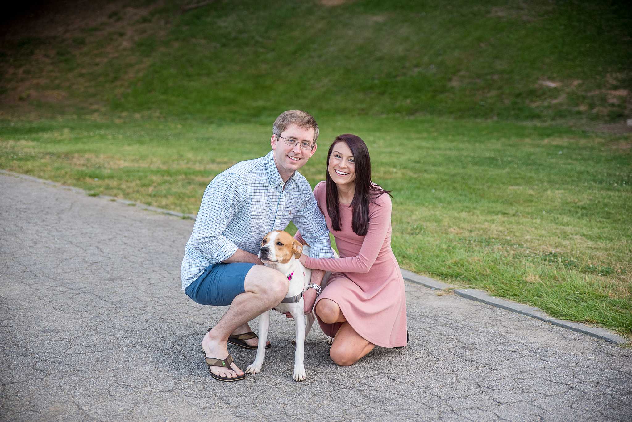 Couple kneeling on a paved path with their dog between them and a green hillside behind at Piedmont Park