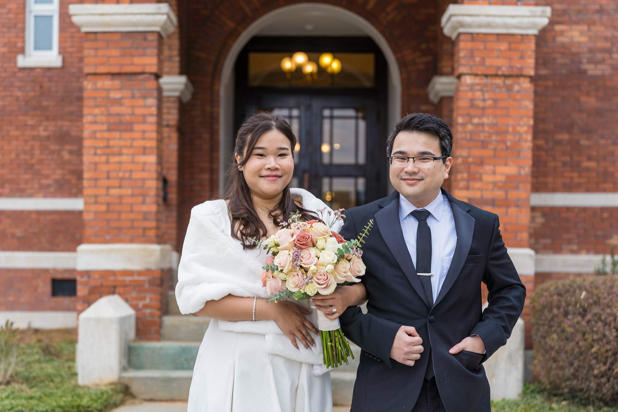 Couple smiling in front of the arched entrance of the Gwinnett Historic Courthouse, the bride wearing a white wrap