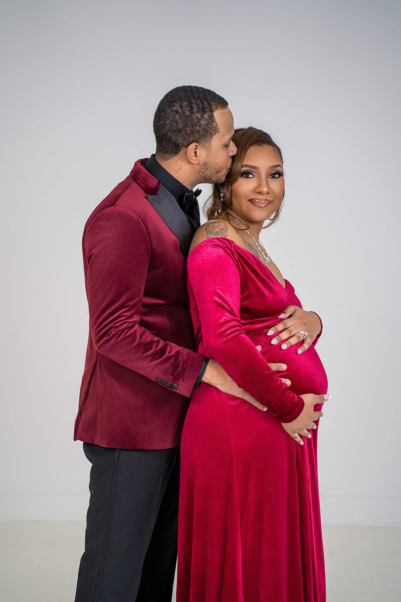 Expecting couple in a red maternity gown and burgundy jacket at White Wall Studio