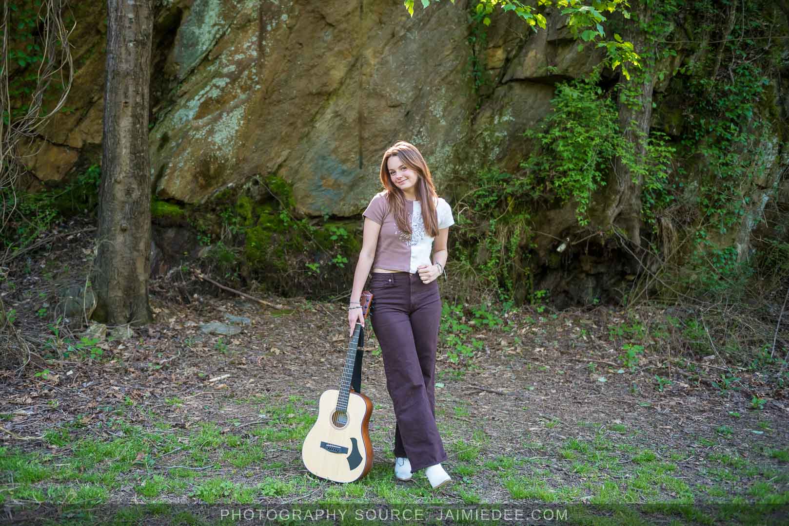 Wide senior portrait with an acoustic guitar beside a rock wall and trees at Olde Rope Mill Park