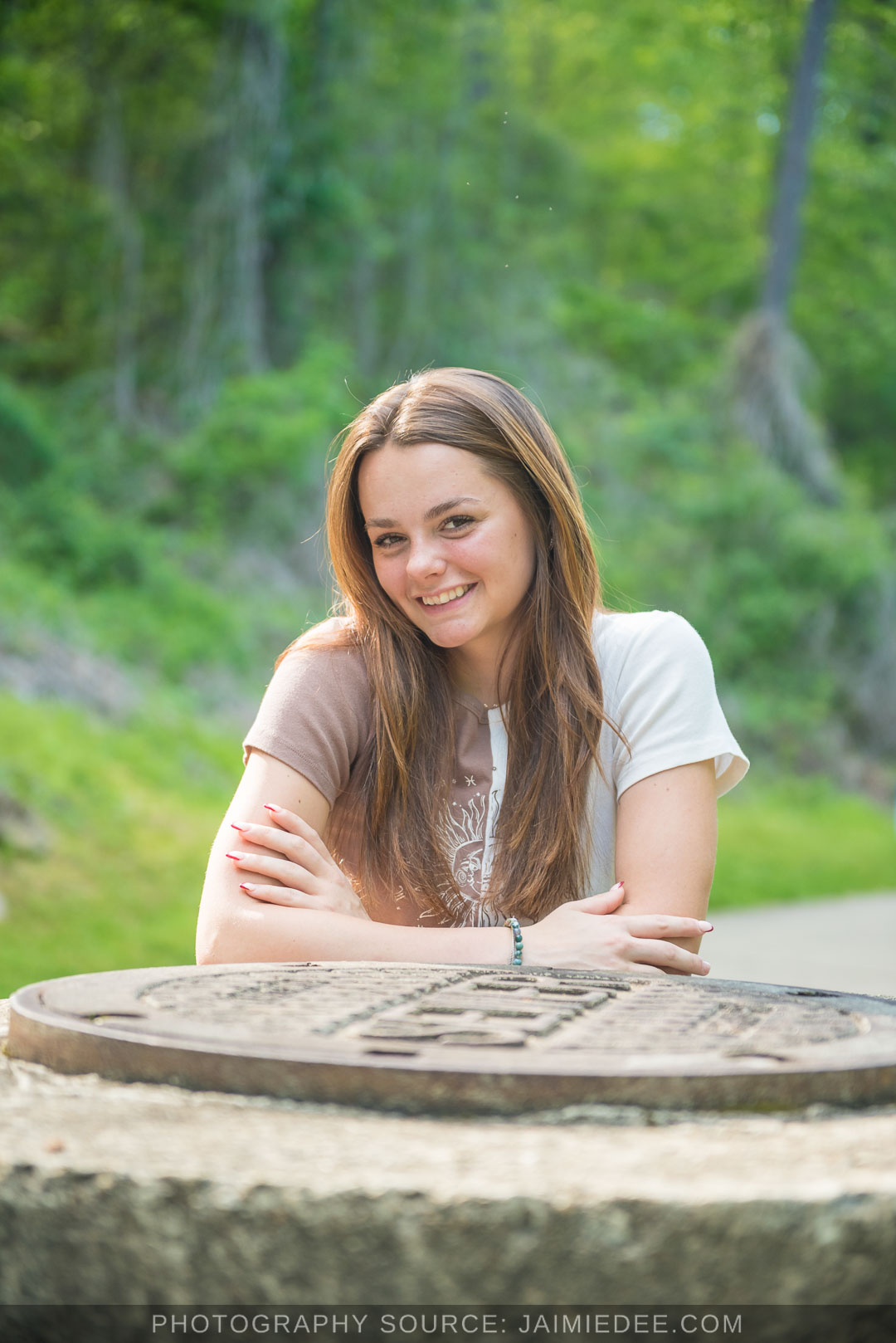Smiling high school senior leaning on a round stone with green trees behind her at Olde Rope Mill Park