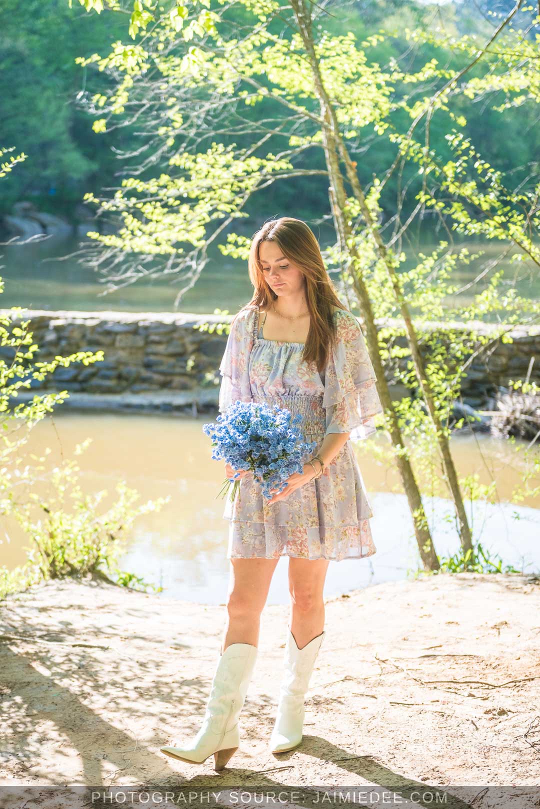 Senior holding blue flowers on the sandy riverbank with the Little River behind her at Olde Rope Mill Park